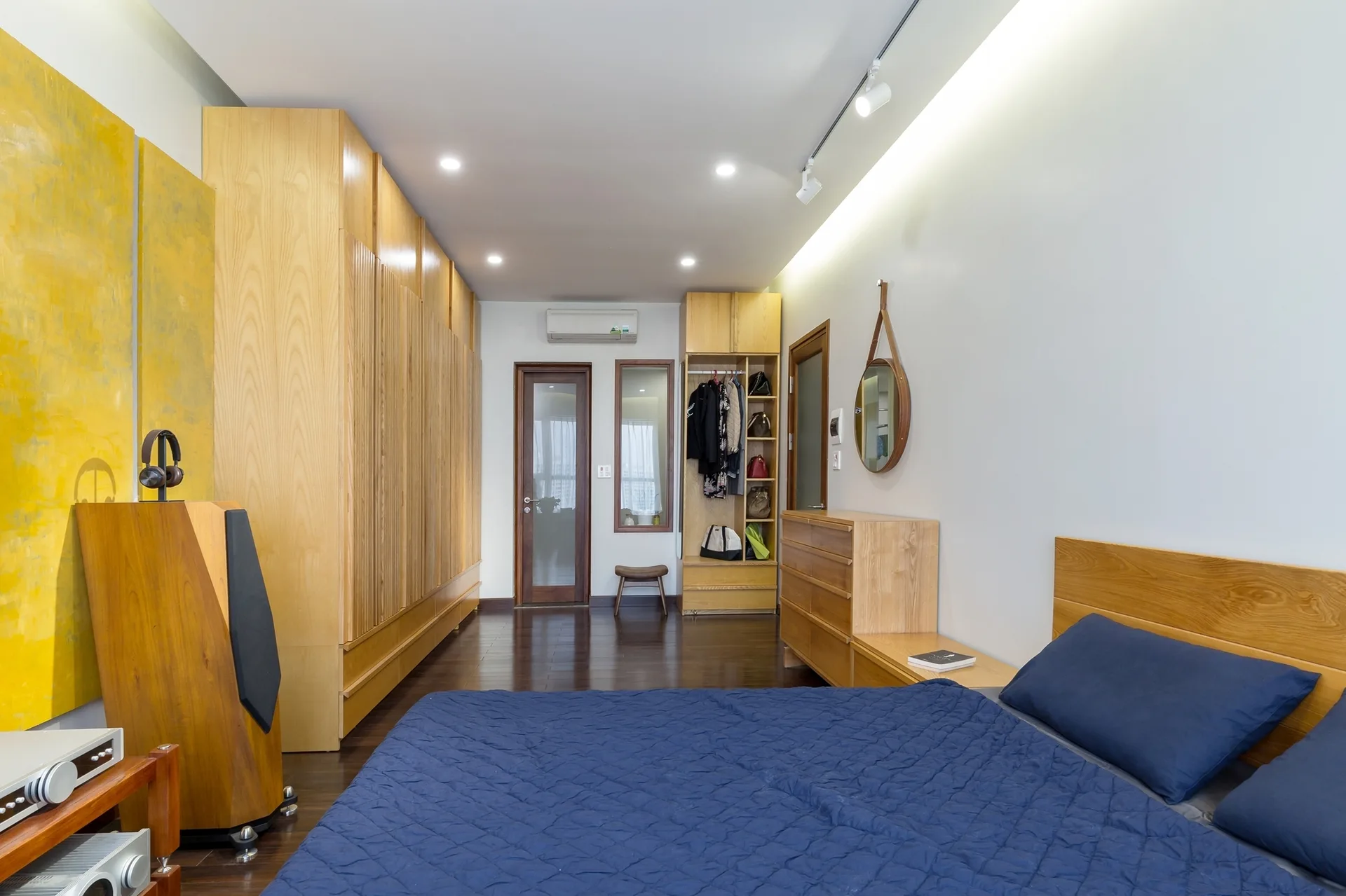 A modern bedroom features custom light wood wardrobes along one wall, dark wood floors, and a bed with blue quilted bedding.