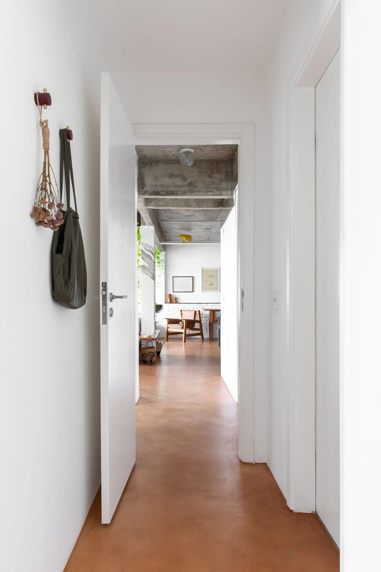 A white-walled hallway with a matte terracotta floor leads to a room with exposed concrete ceiling beams and a wooden armchair.