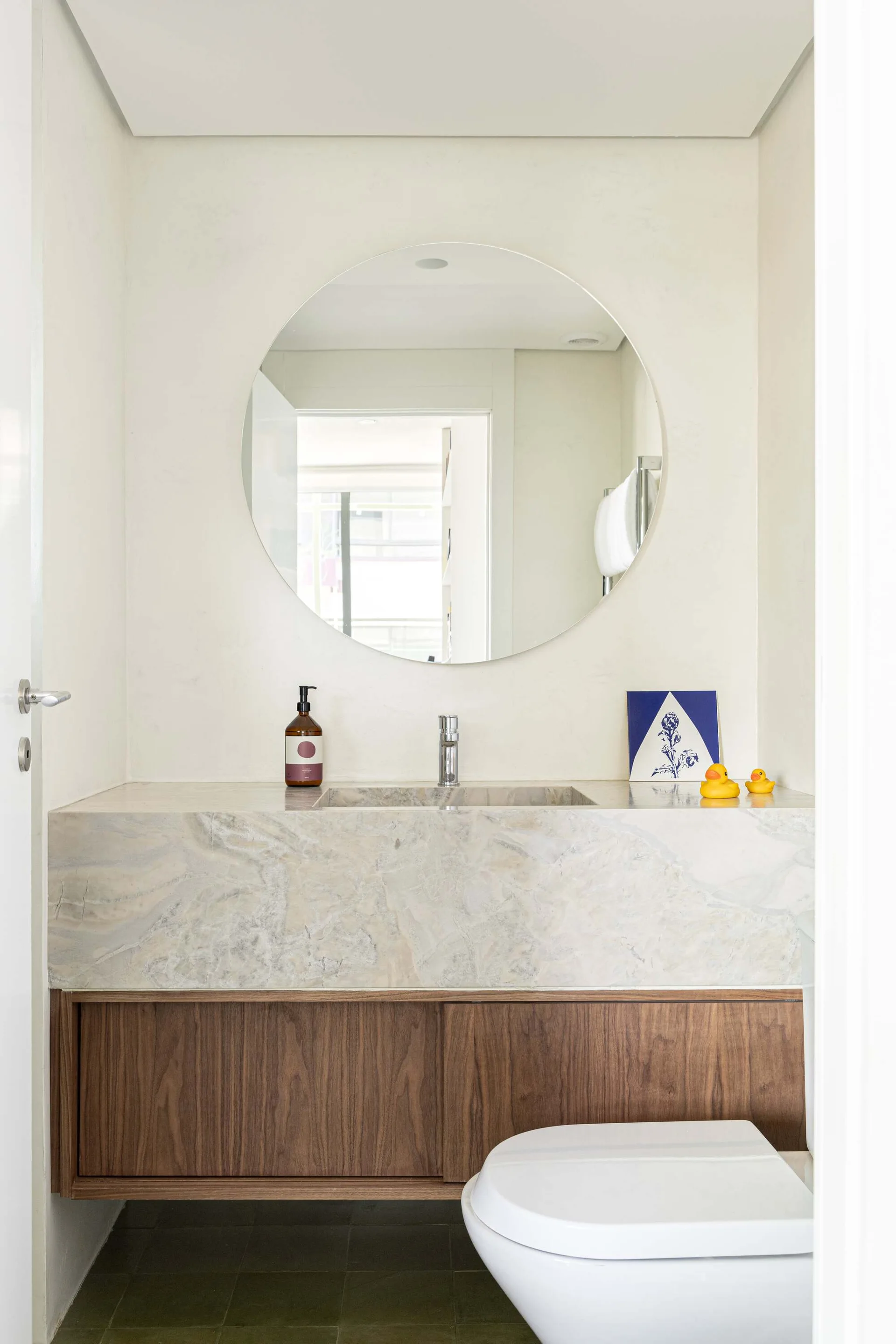 A compact powder room contains a floating vanity with a marble countertop, wood cabinet, a large round mirror, and a white toilet.