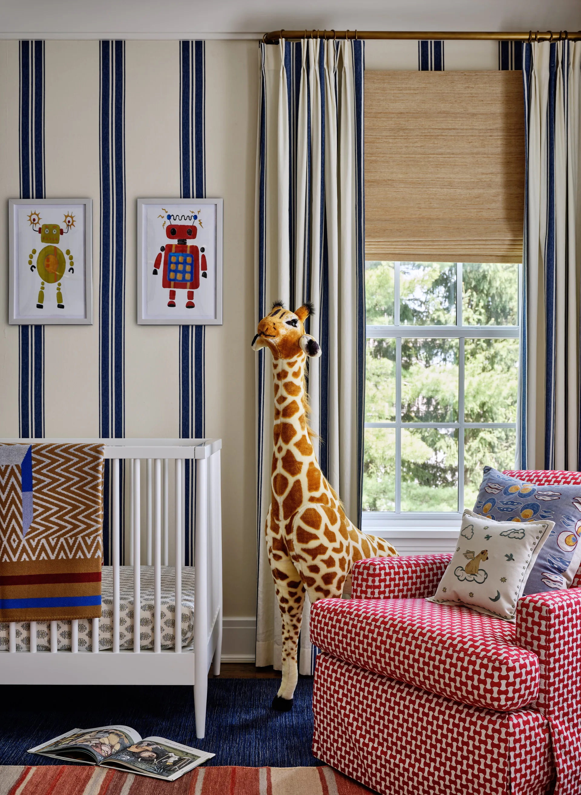 A bright nursery features a white wooden crib, a tall plush giraffe toy, and a red patterned armchair against blue and beige striped wallpaper.