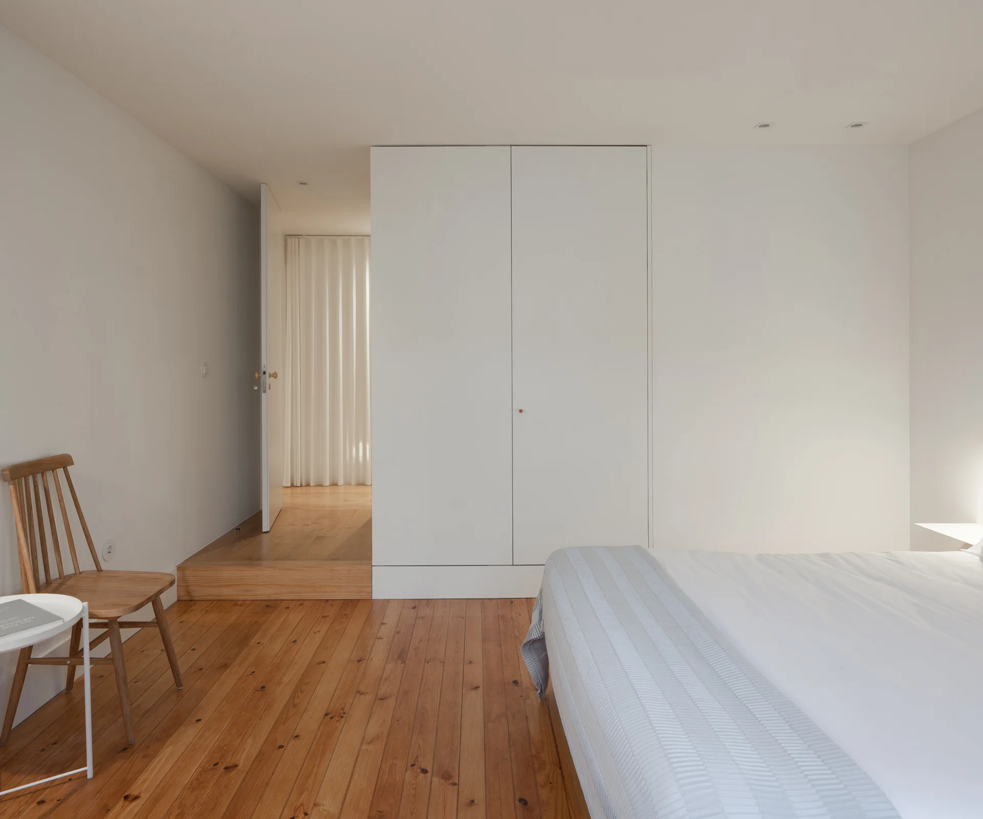 A minimalist bedroom with rich wood plank flooring, a white built-in wardrobe, a wooden spindle-back chair, and a bed with light-colored bedding.