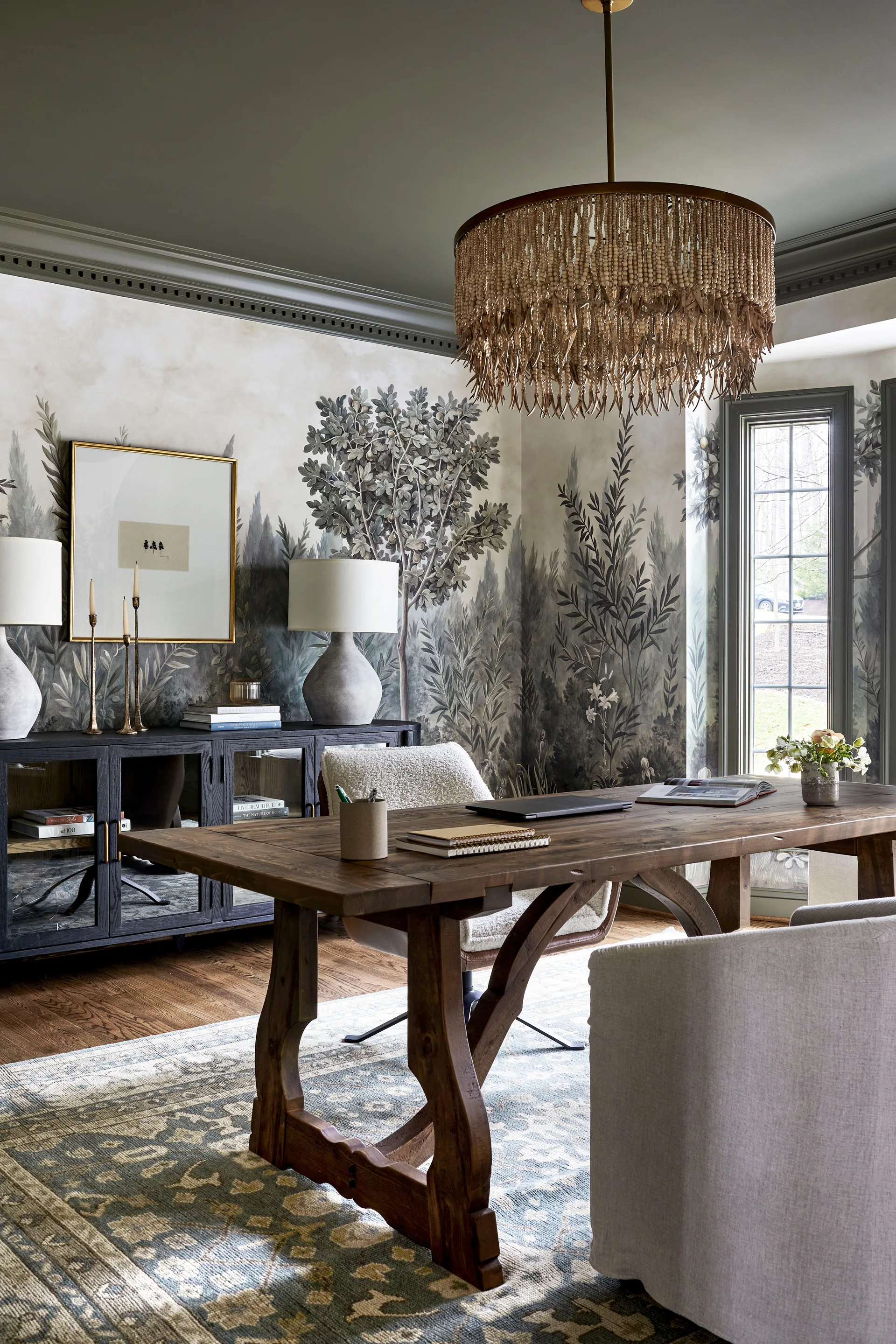 A home office features a rustic wooden trestle desk, a white bouclé office chair, botanical mural wallpaper, and a beaded pendant light.