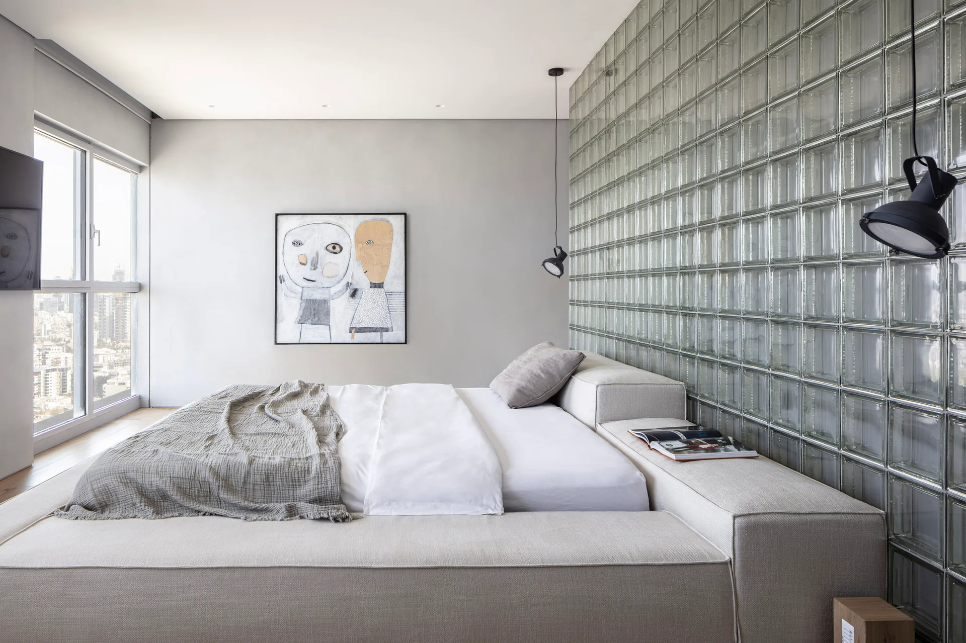 A contemporary bedroom features a low-profile upholstered bed next to a large glass block wall, with a minimalist framed art piece on the back wall.