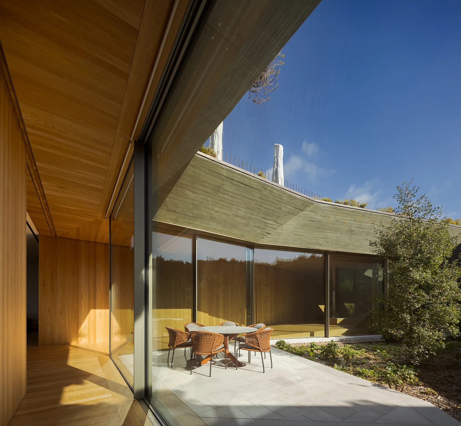 A modern patio features a round dining table with woven chairs next to large glass walls that reveal a warm wood-clad interior.