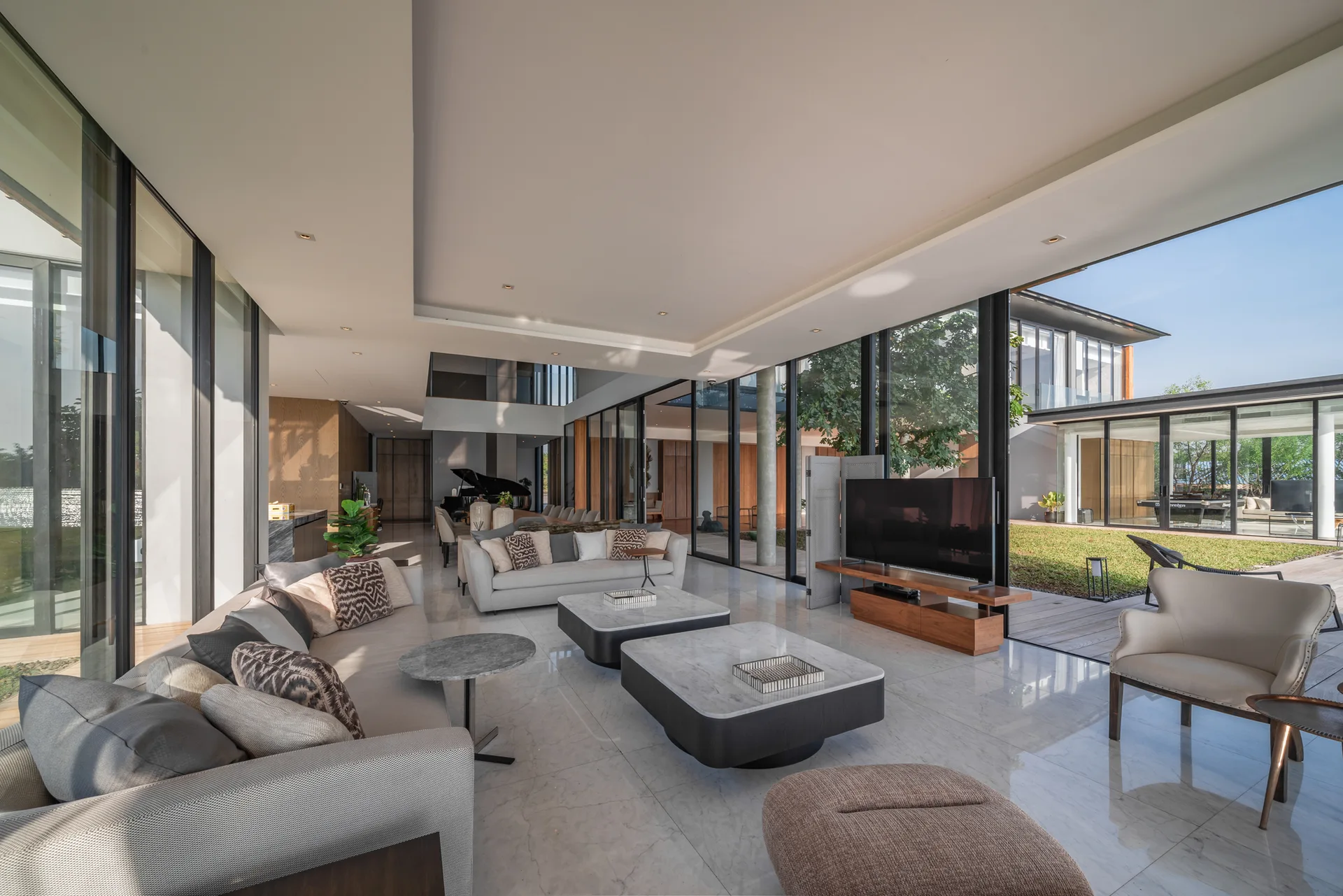 A spacious modern living room features floor-to-ceiling glass walls overlooking a courtyard, neutral sofas, and polished stone flooring.