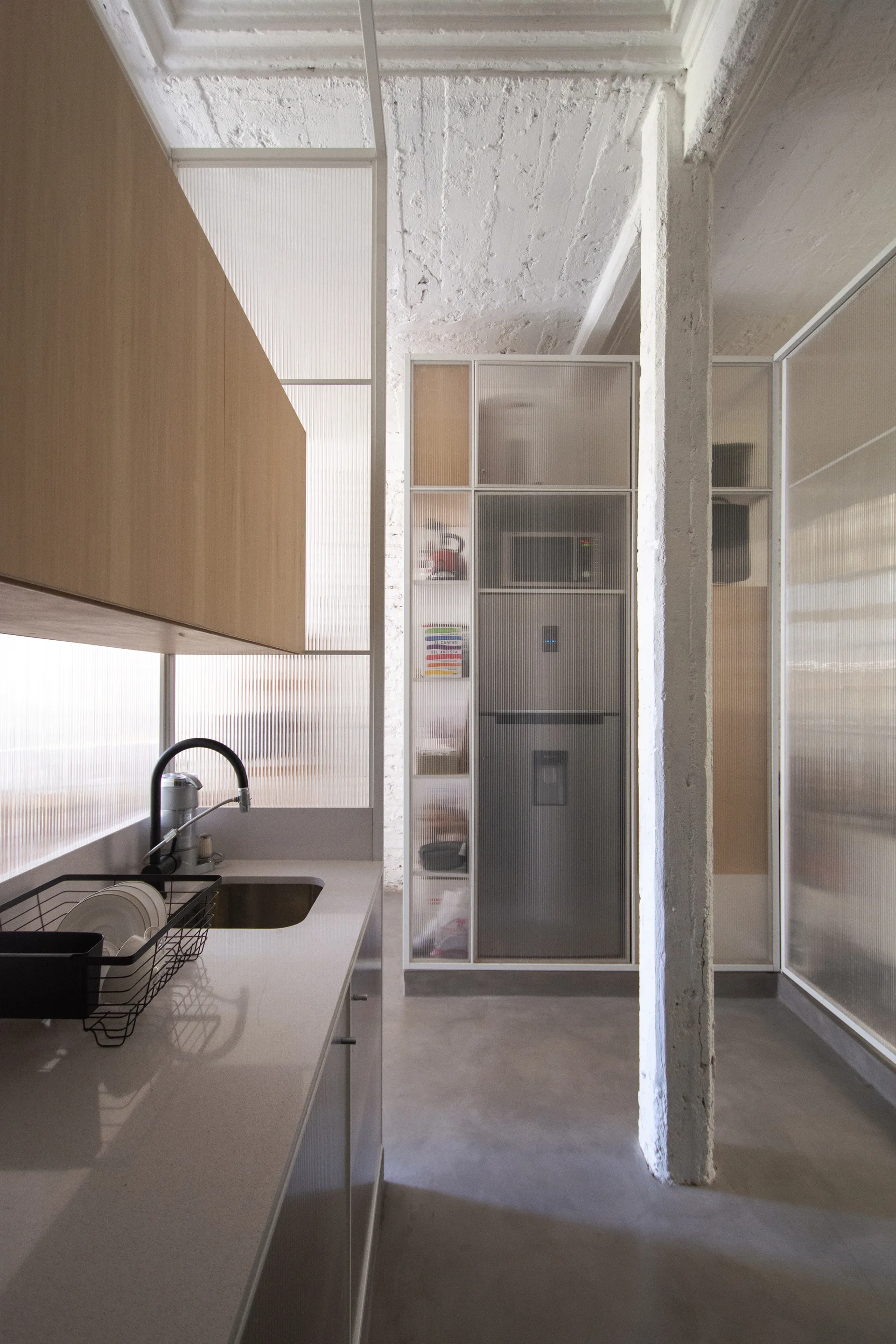 A modern industrial kitchen with a grey concrete floor, a textured white structural column, light wood cabinetry, and ribbed glass partition walls.