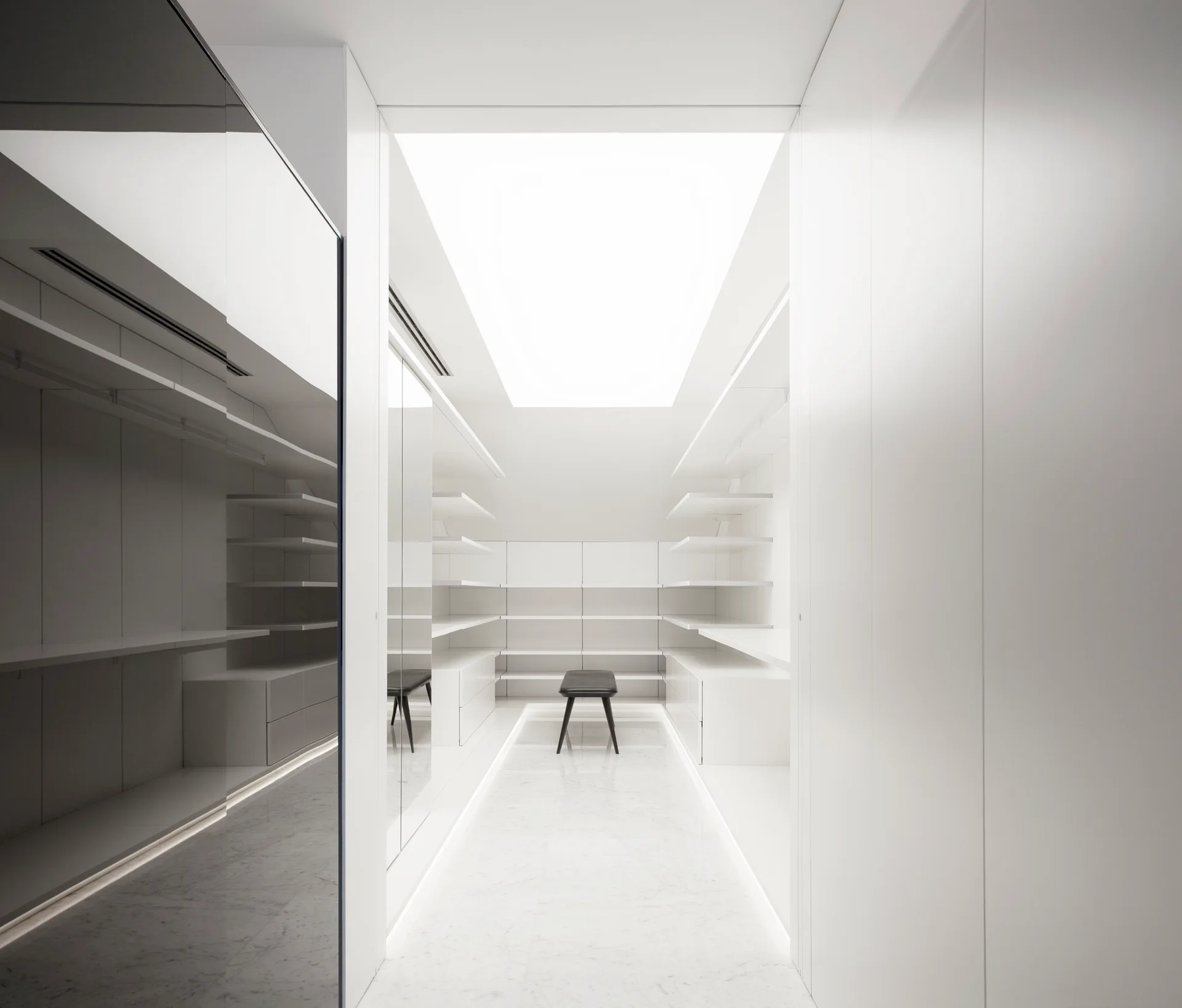 A minimalist white walk-in closet featuring symmetrical open shelving, a large central skylight, polished stone floors, and a single black stool.