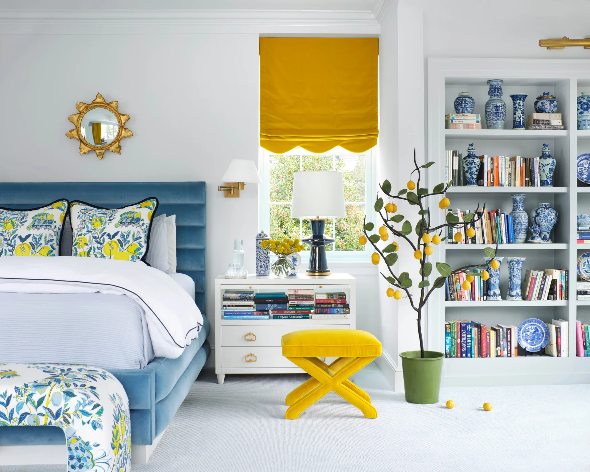 A bedroom features a blue velvet bed, a yellow scalloped roman shade, a yellow x-shaped stool, and a white built-in bookshelf.