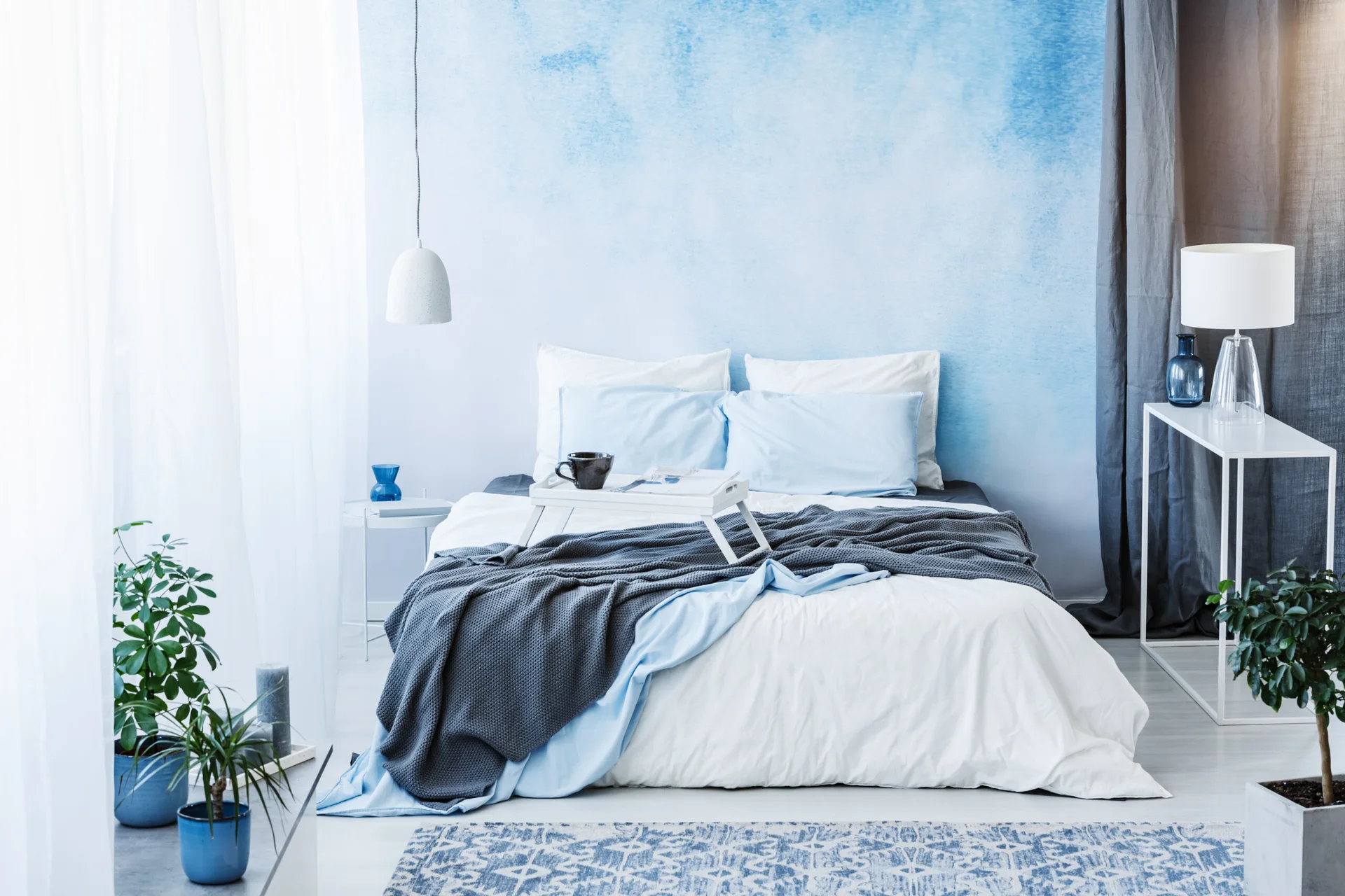 A modern bedroom features a bed with blue and white linens against a blue watercolor ombre accent wall, flanked by sheer curtains and simple side tables.