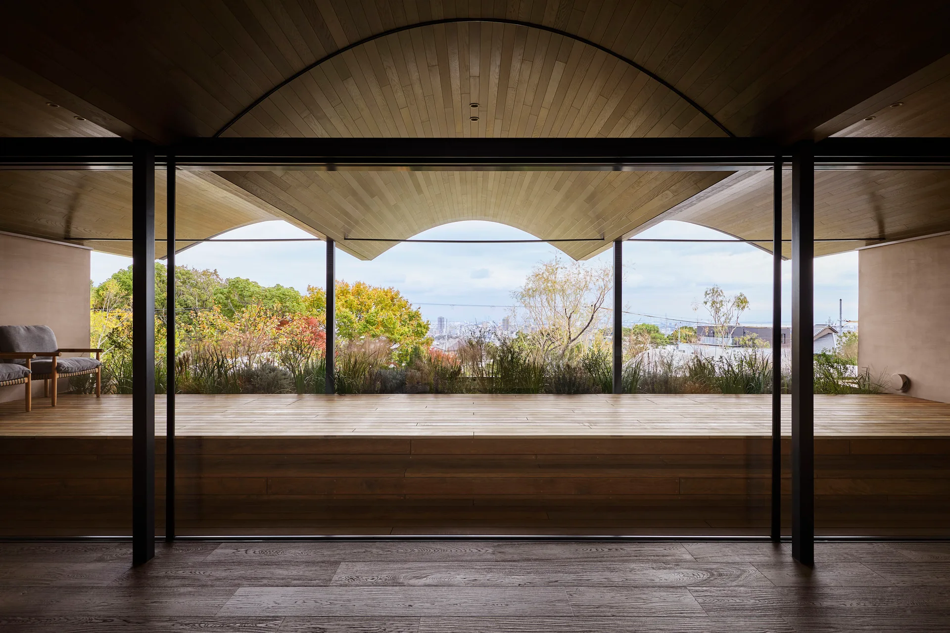 A covered wooden terrace with a curved wood paneled ceiling features a large glass wall looking out over a landscape with green trees.