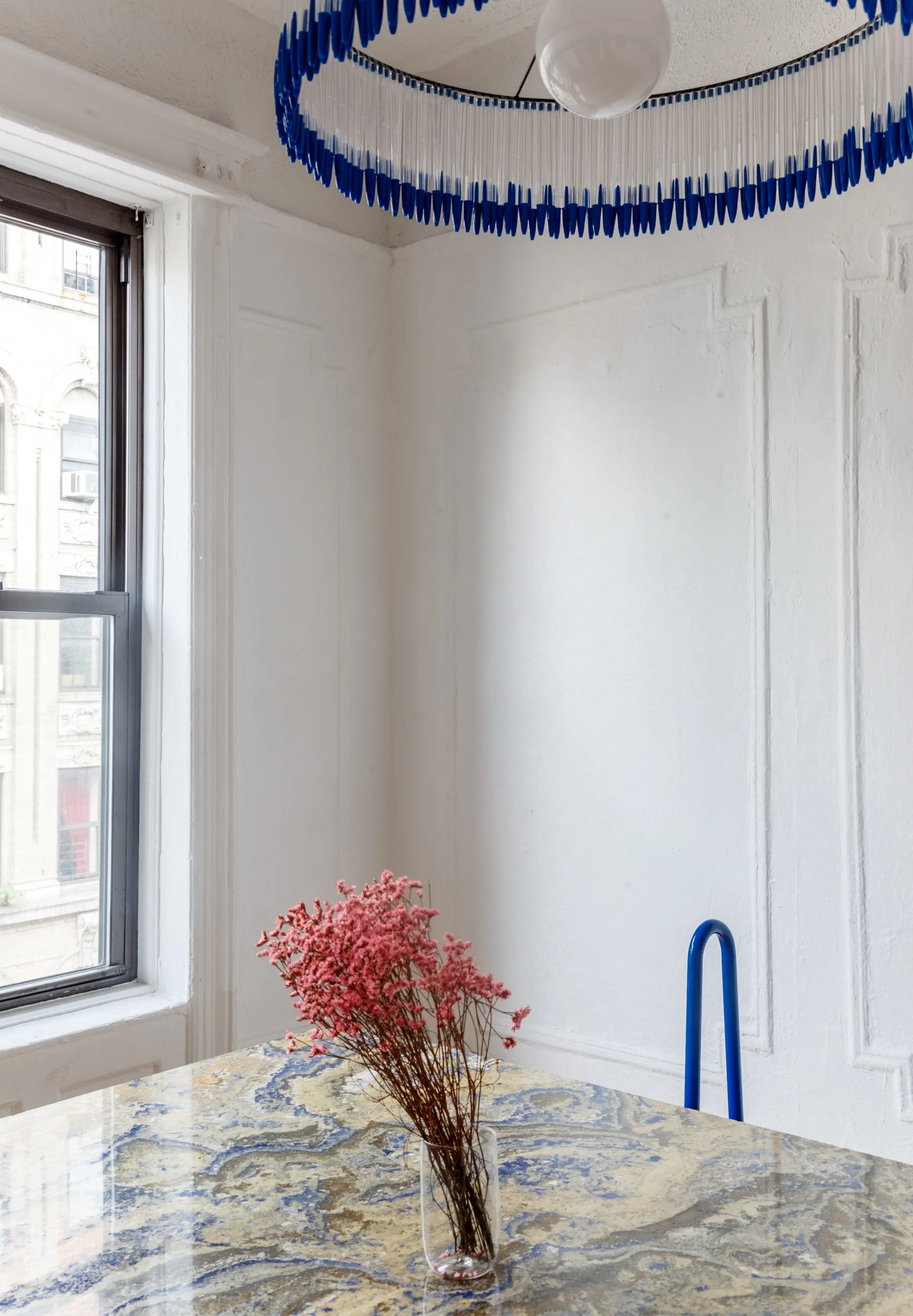 A dining space features a blue and gold marble table, a cobalt blue chair frame, and a fringed blue pendant light against white paneled walls.