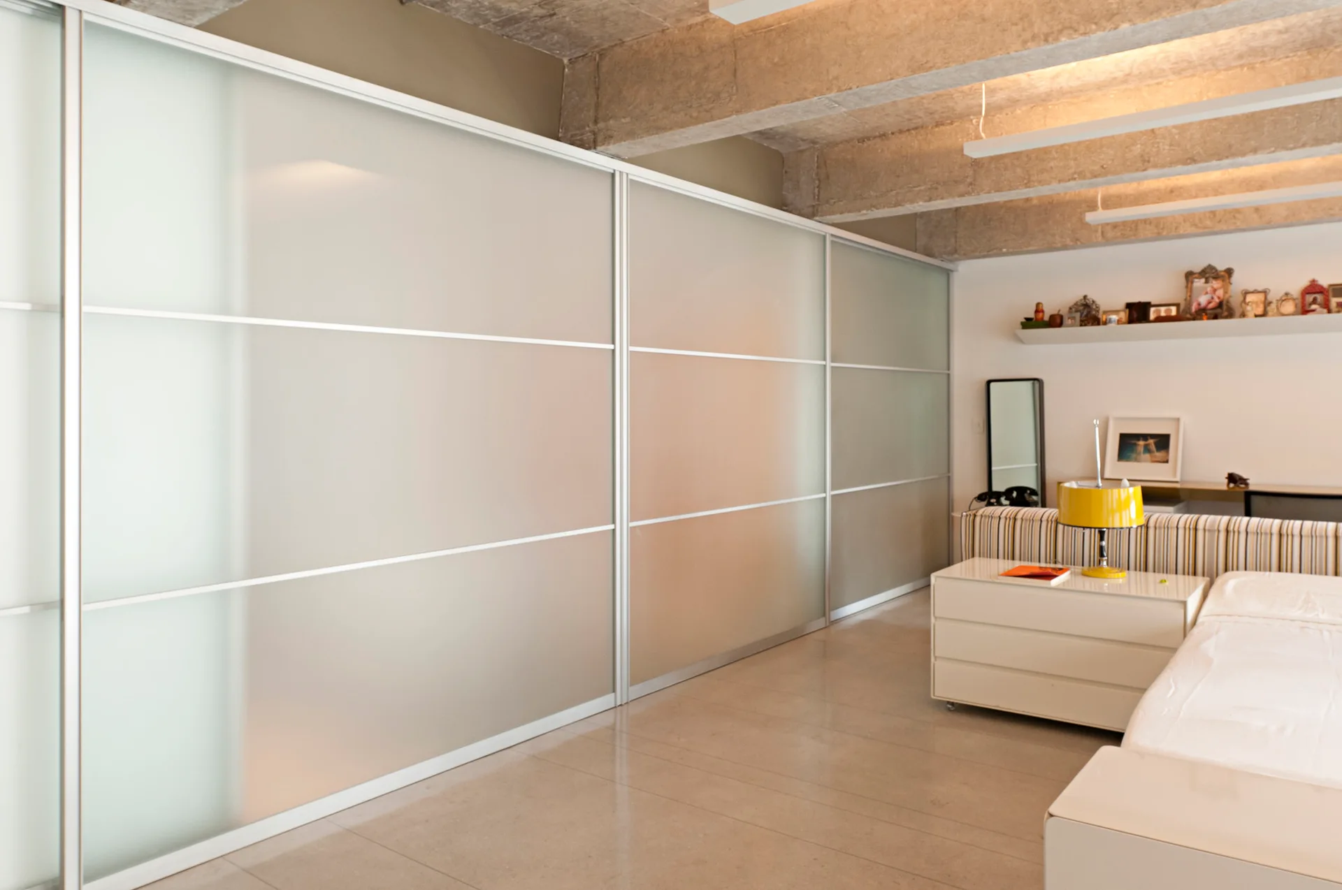 A minimalist bedroom features large frosted glass sliding partition doors beneath exposed concrete ceiling beams and a white nightstand with a yellow lamp.