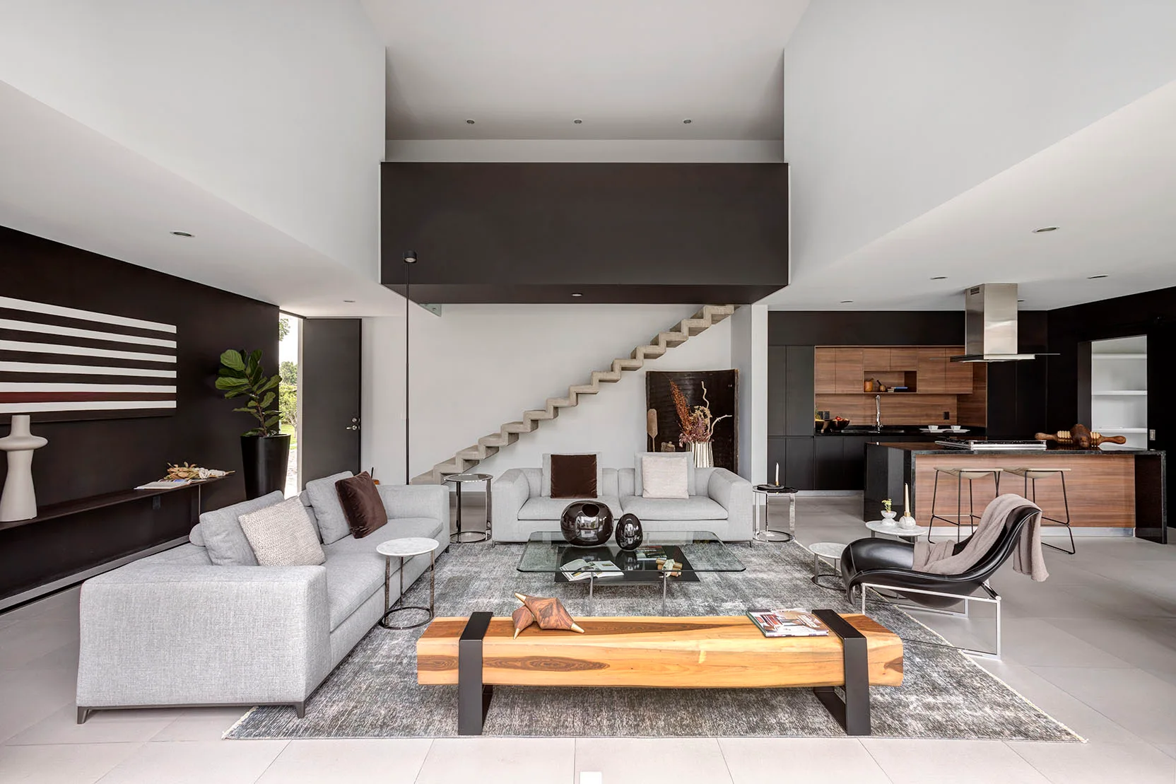 A modern open-concept living room and kitchen featuring two gray sofas, a wood block bench, and a concrete floating staircase against a white wall.