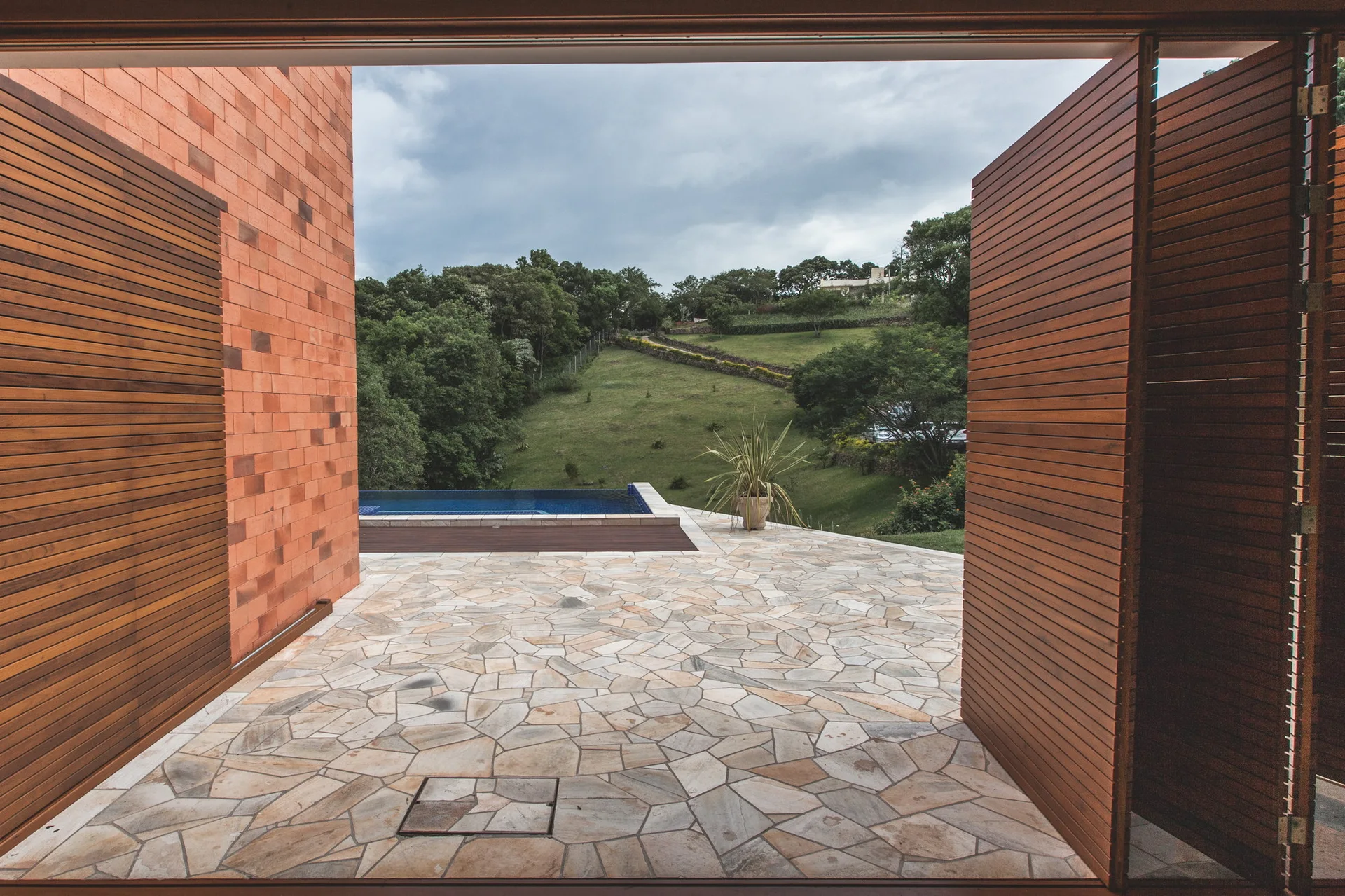 A view looking out through open slatted wood doors onto a flagstone patio with a swimming pool and grassy hills in the distance.