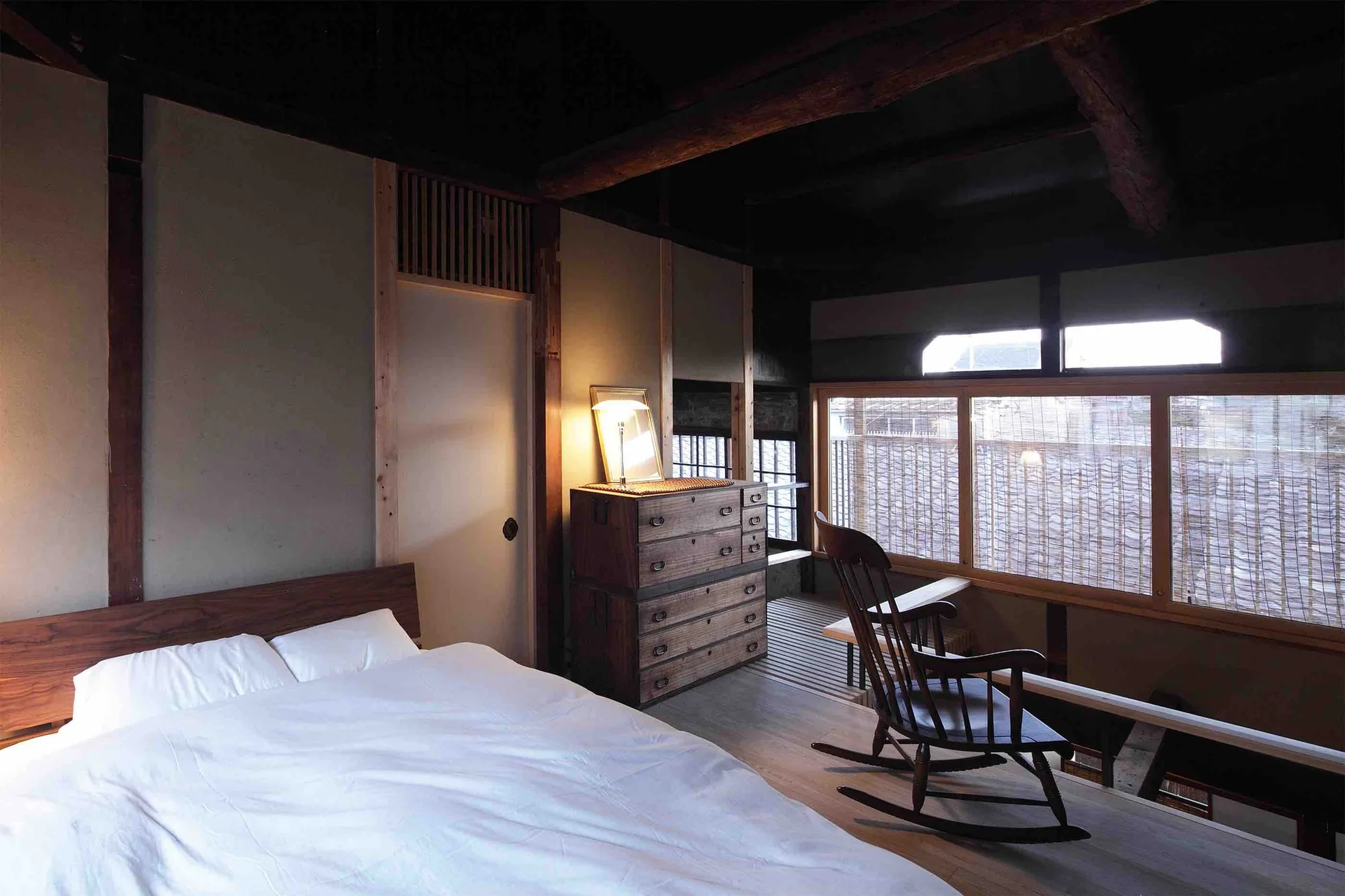 A minimalist bedroom featuring a low-profile bed, an antique wooden chest of drawers, a rocking chair, and dark exposed timber ceiling beams.