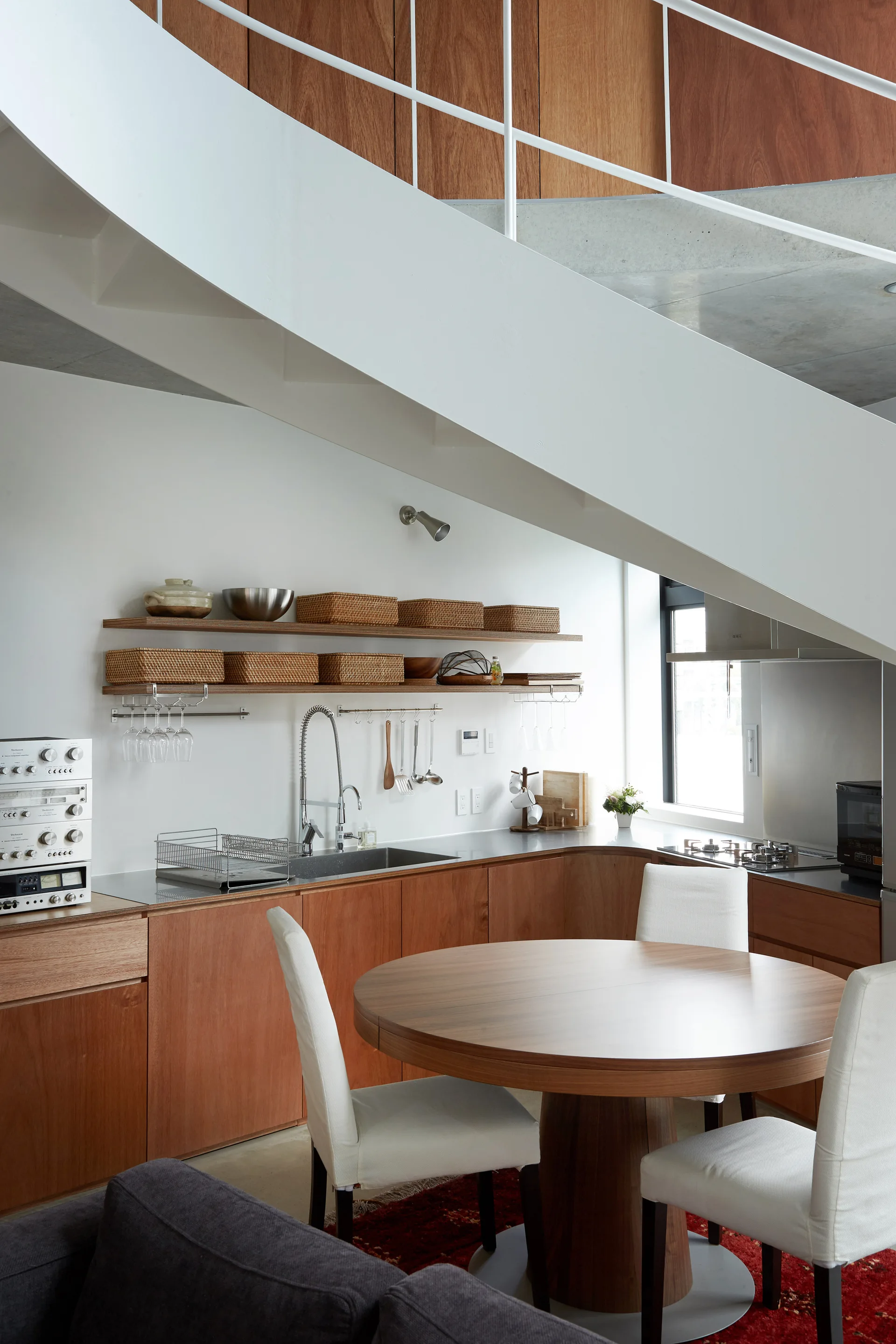 A wood-paneled kitchen features a round dining table with white chairs beneath a diagonal white staircase and wooden open shelving.