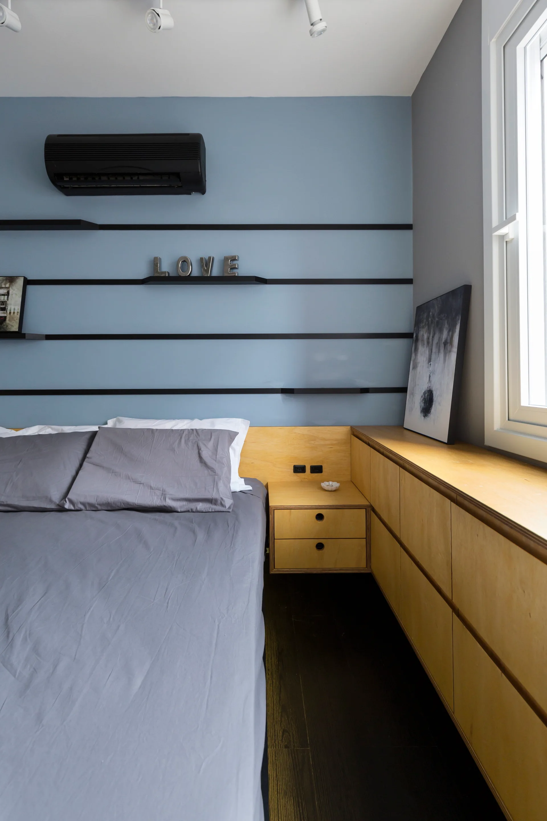 A modern bedroom features a gray bed, integrated light wood cabinetry, and a blue accent wall with thin black horizontal shelves.