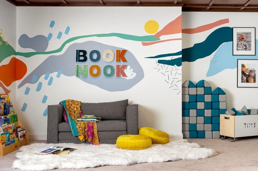 A kids' playroom features a grey sofa, yellow cushions, a white faux-fur rug, and a colorful abstract wall mural spelling out BOOK NOOK.