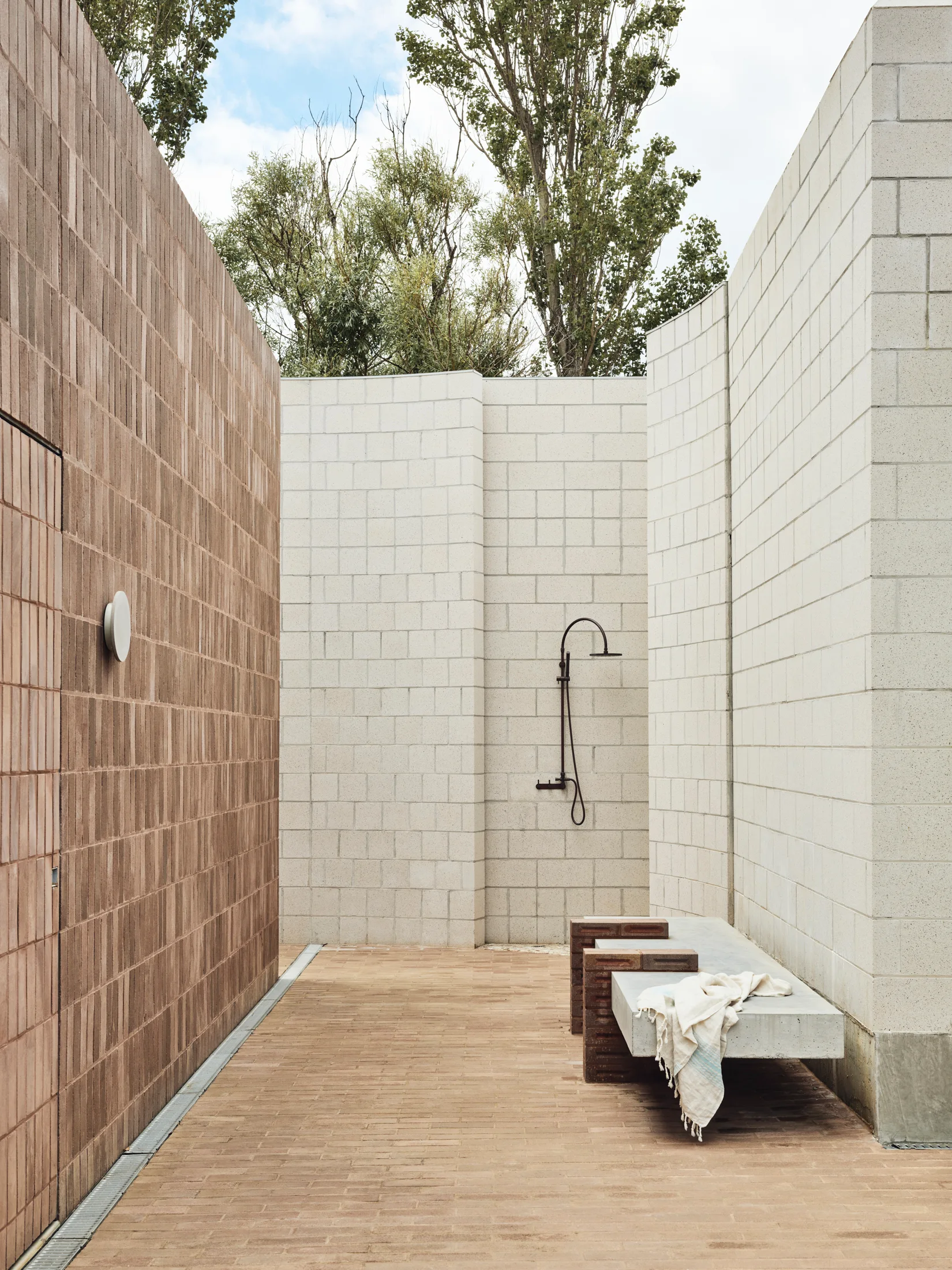 An outdoor shower area with tall off-white masonry walls, a black metal shower fixture, a concrete bench, and a terracotta brick floor.