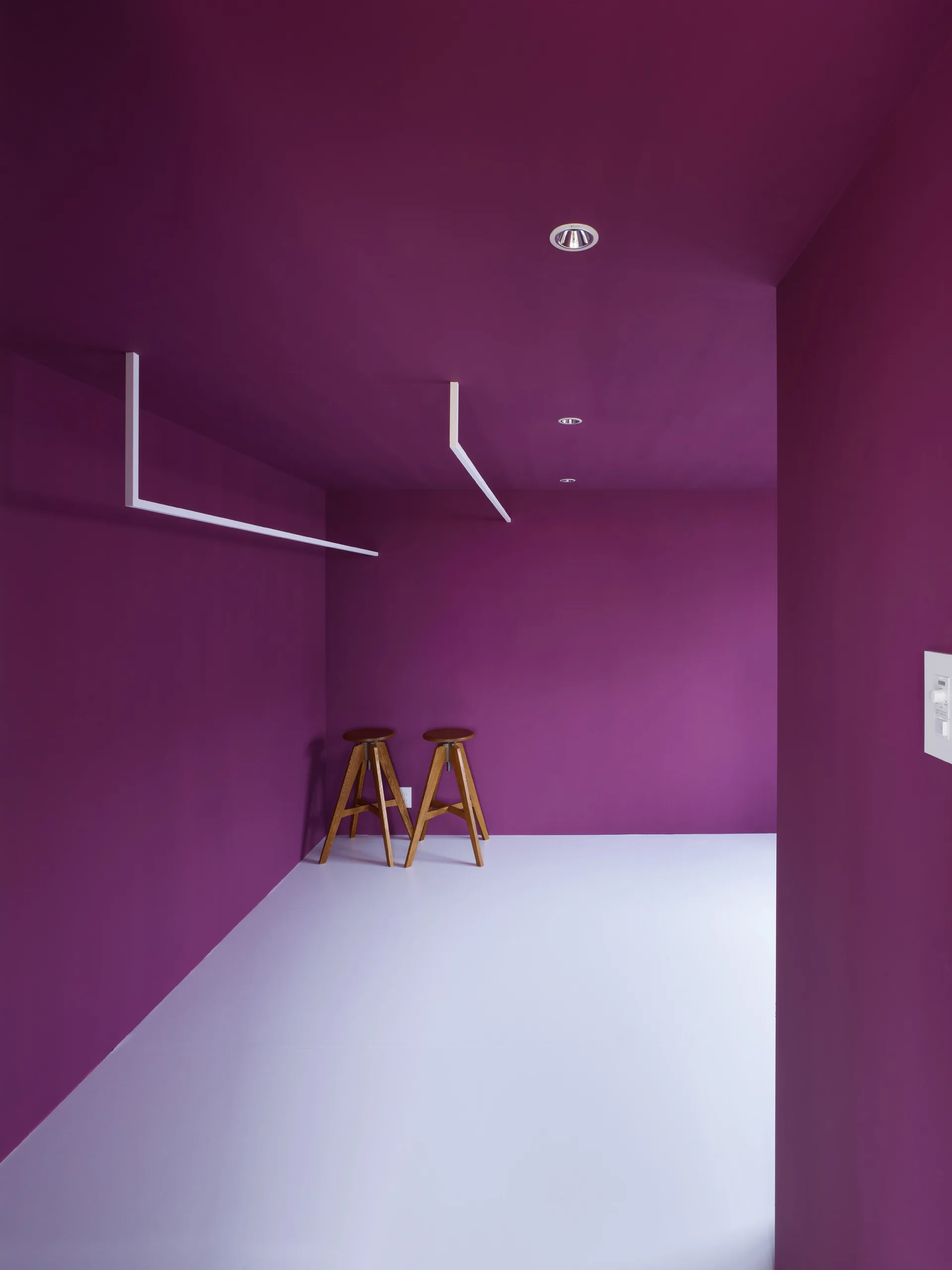 Two wooden stools stand in the corner of a minimalist room with bold magenta painted walls and ceiling and a solid white floor.