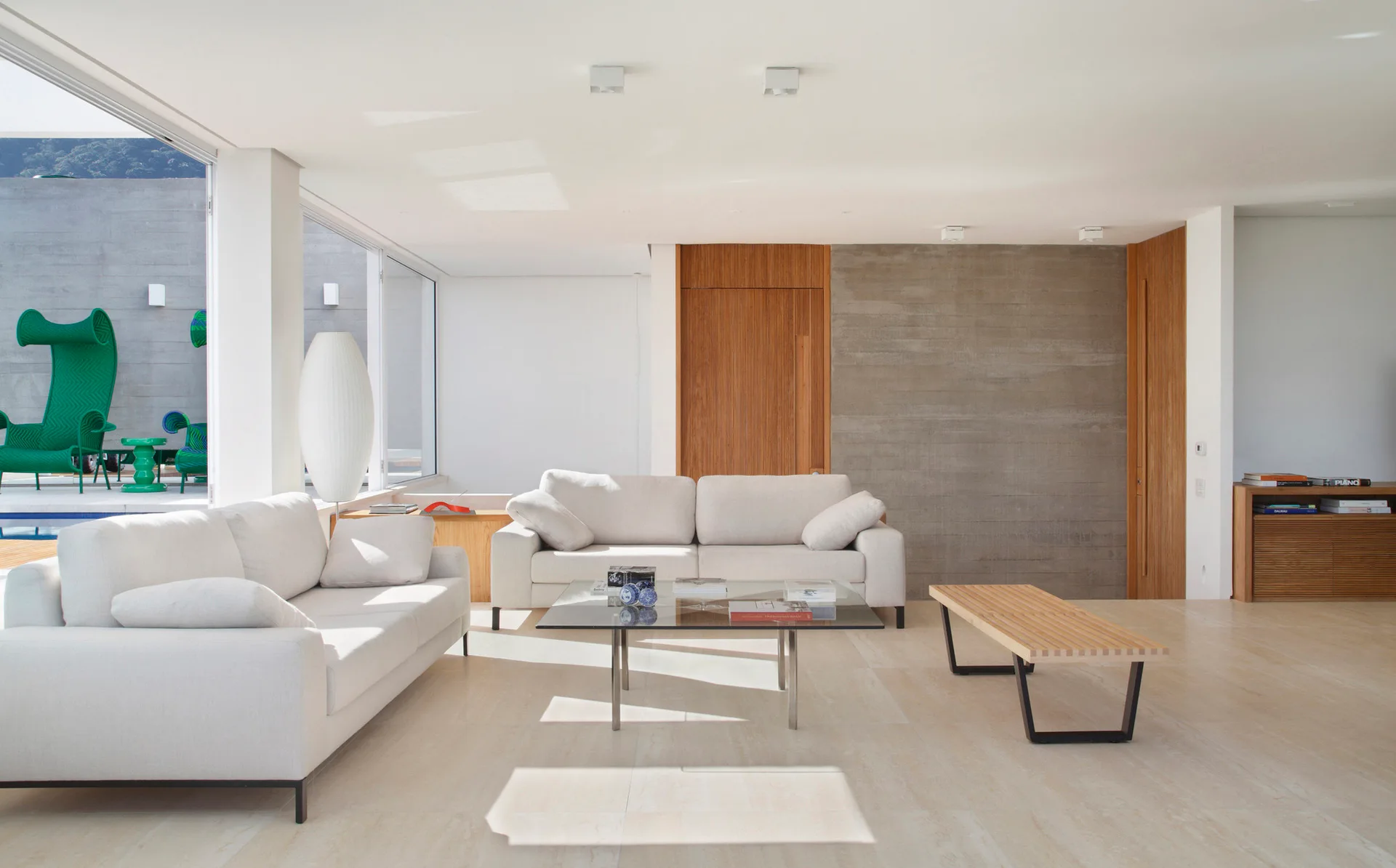A bright minimalist living room features two off-white sofas, a glass-top coffee table, and an accent wall combining raw concrete and warm wood paneling.