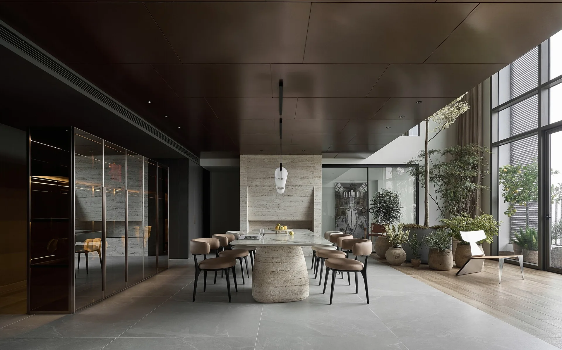 A minimalist dining room with a stone table, curved chairs, a dark paneled ceiling, a stone fireplace backdrop, and large windows with potted plants.