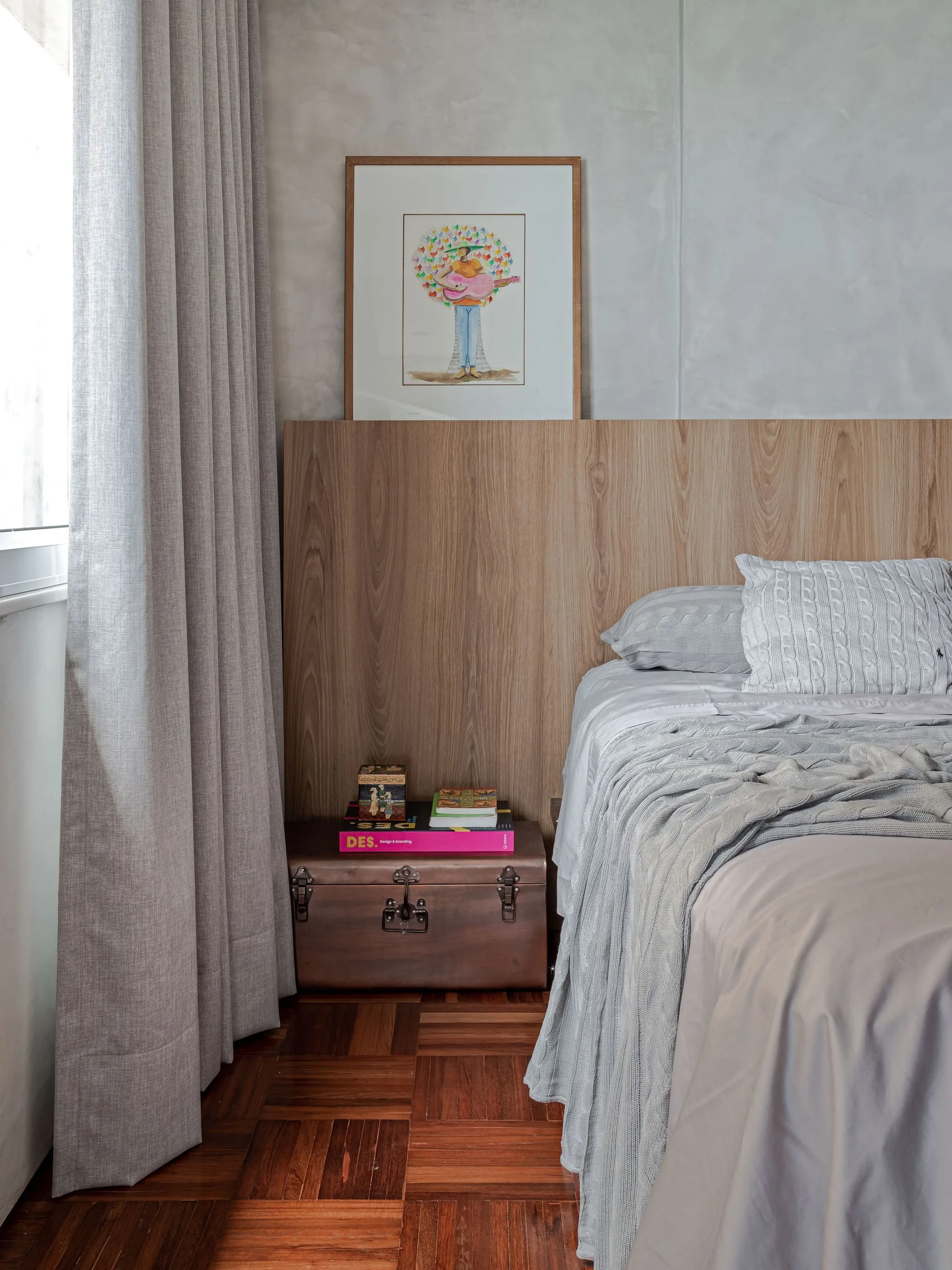 A bedroom corner featuring a wood panel headboard, a vintage trunk nightstand, and grey bedding on a parquet floor.