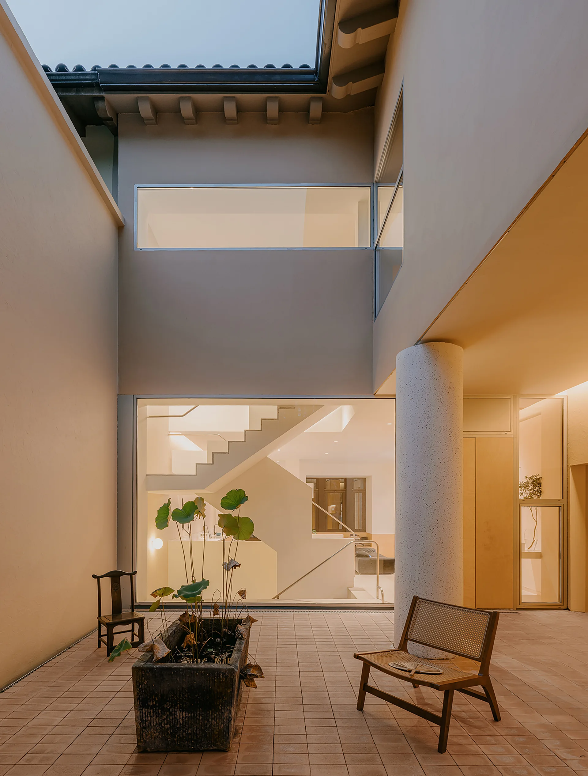 An outdoor paved courtyard features a rectangular stone planter, a cane armchair, and a large window showcasing a warmly lit interior staircase.