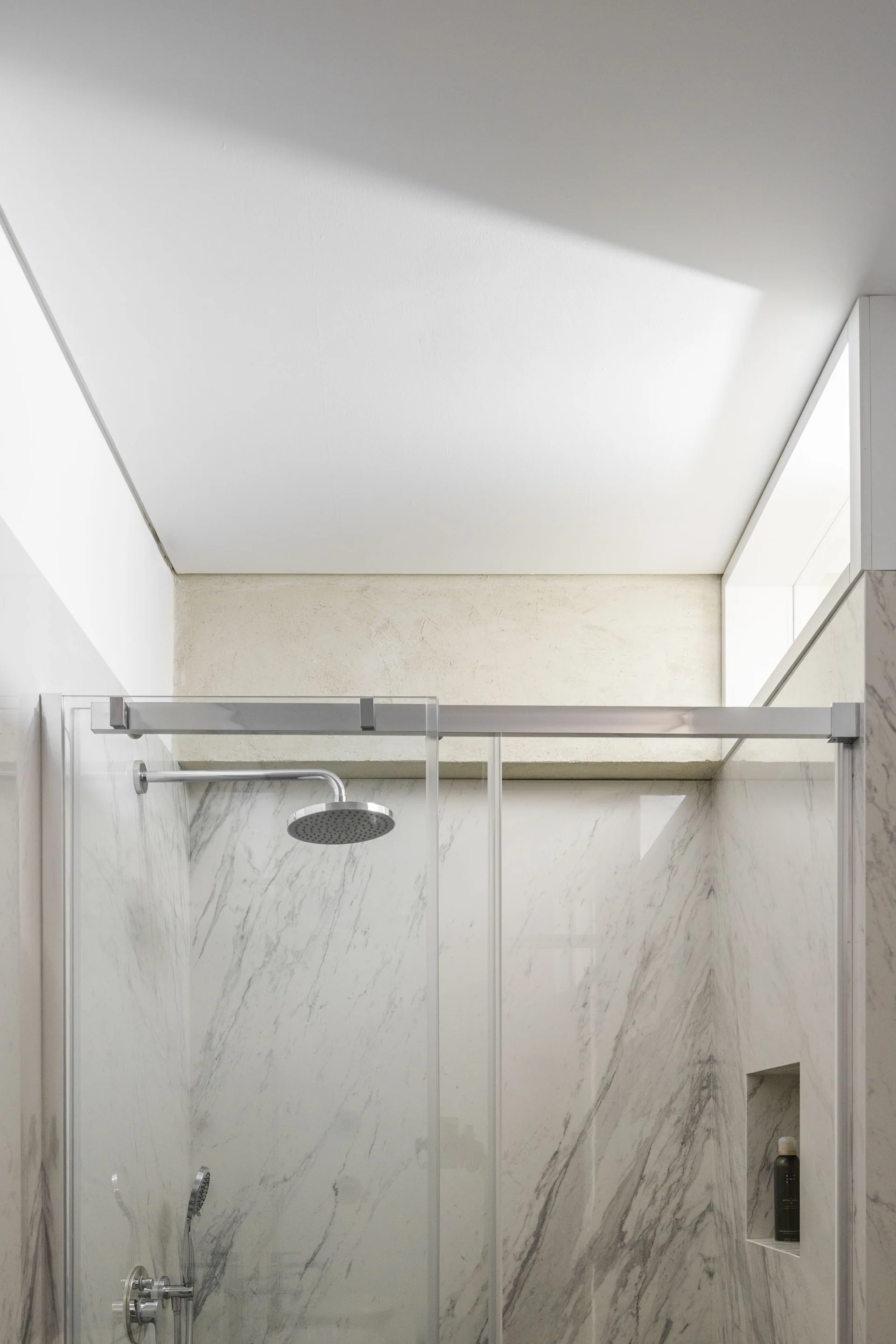 A modern bathroom shower features veined white marble walls, a sliding glass door, a rainfall shower head, and a recessed wall niche.
