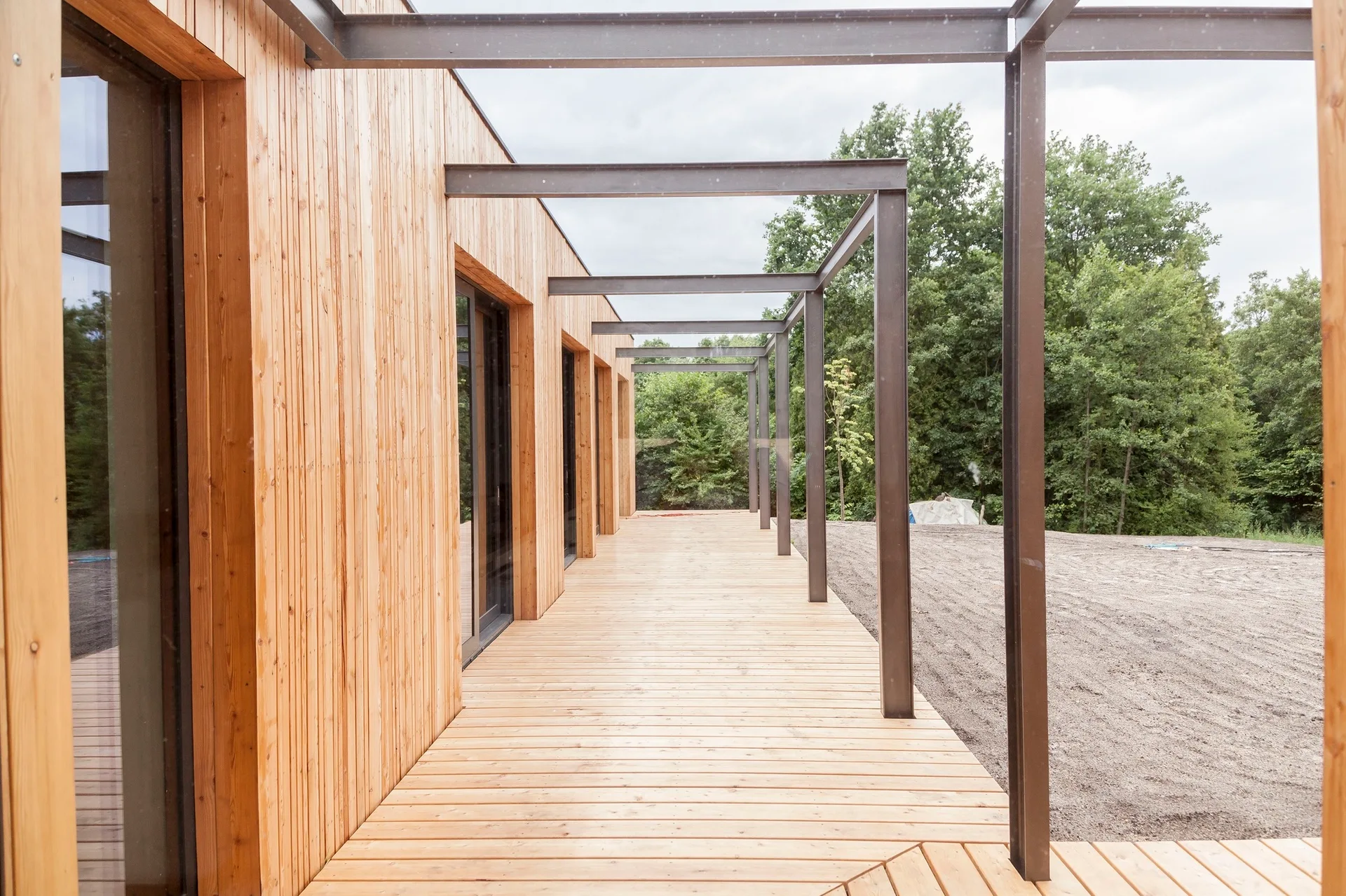 A long wooden deck terrace with light brown vertical wood siding and a dark grey metal pergola structure leading into a forest view.