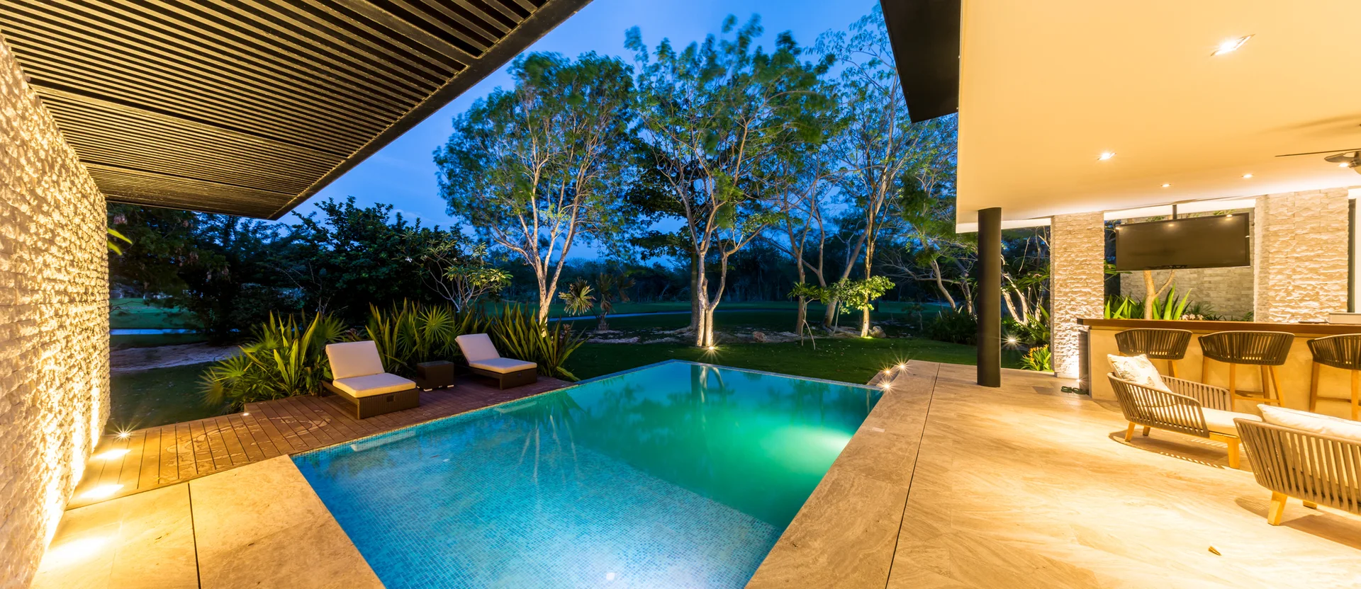 A modern outdoor pool patio at dusk features a turquoise swimming pool, sun loungers on a wood deck, and a stone-clad bar area under a covered ceiling.