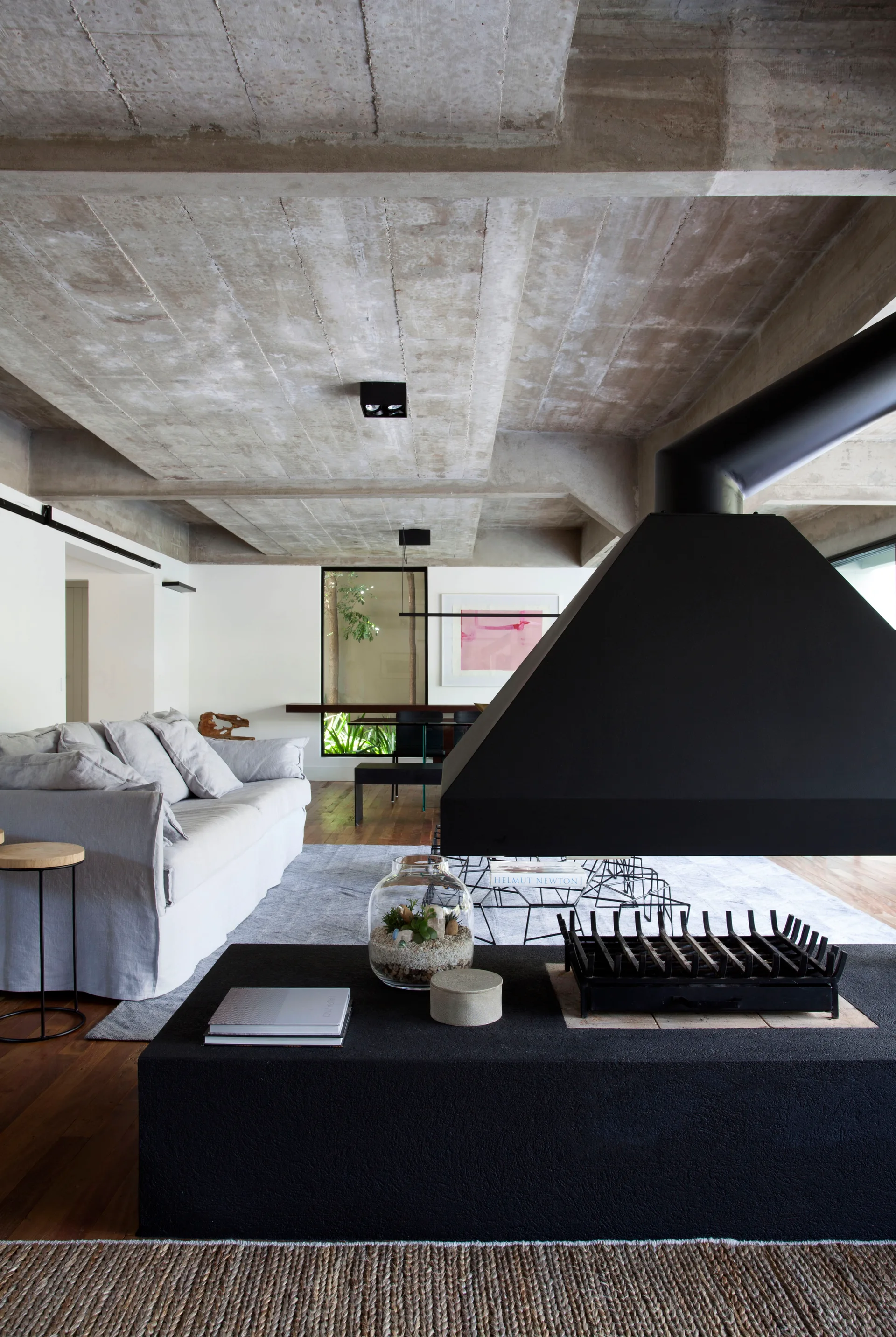 An industrial living room features a grey slipcovered sofa, a black suspended metal fireplace, and exposed concrete ceiling beams.
