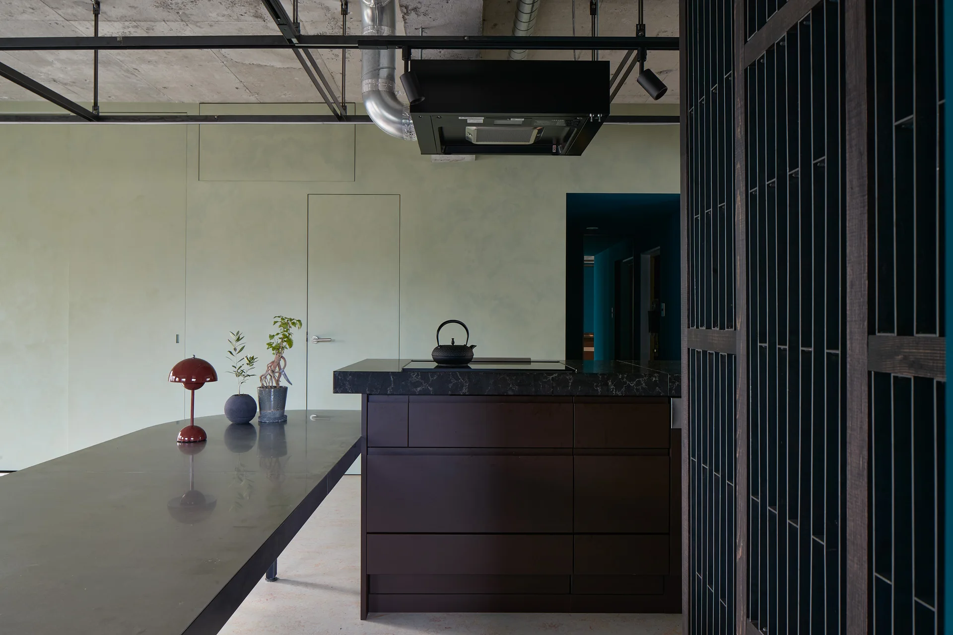 A modern industrial kitchen with a dark stone island, sage green walls, an integrated flush door, and an exposed concrete ceiling with black track lights.
