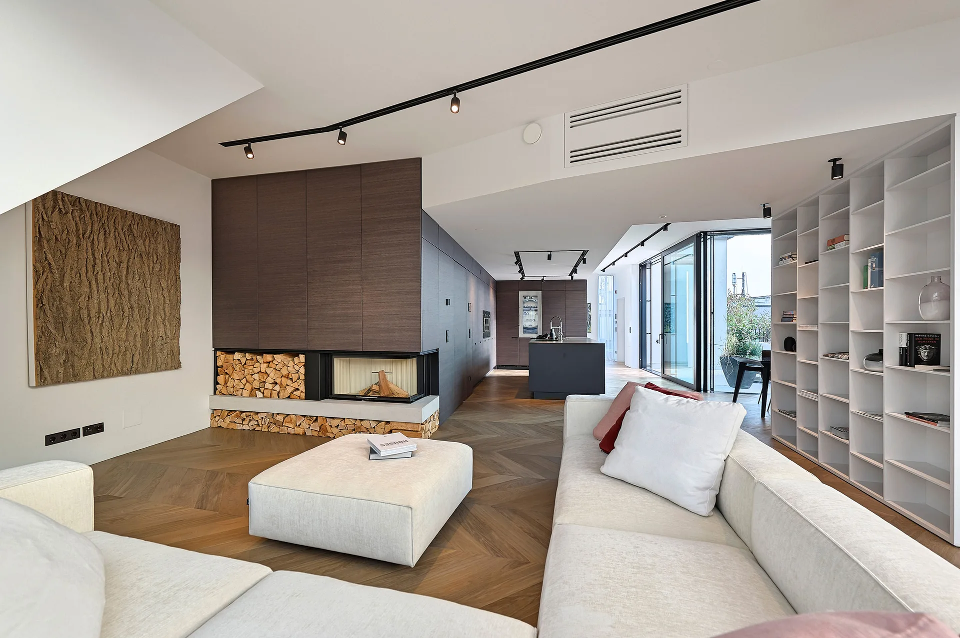 A modern open-concept living room features a cream sectional sofa, chevron pattern wood flooring, and an integrated fireplace with stacked firewood.