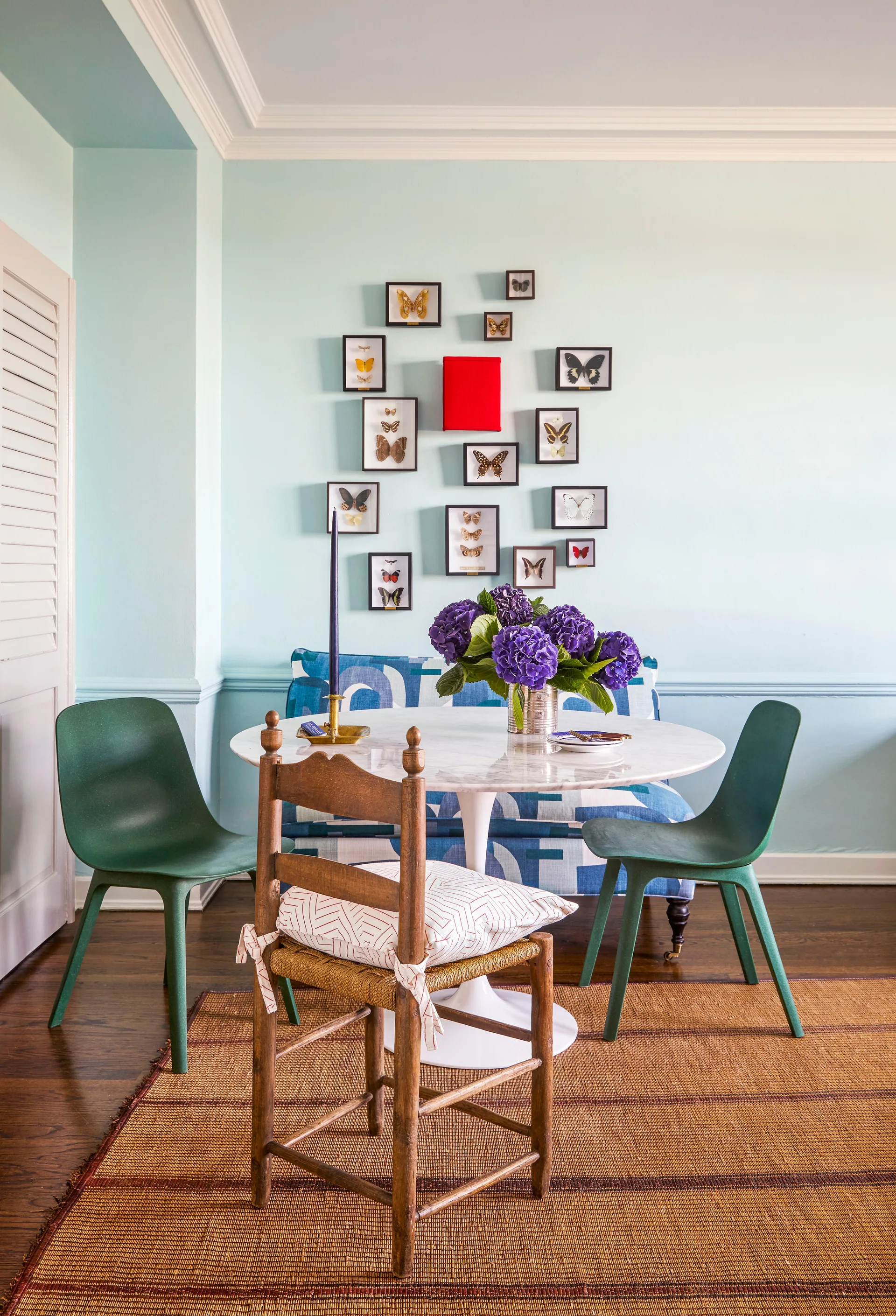 A dining nook with a marble tulip table, blue patterned bench, green plastic chairs, and framed butterfly art on light blue walls.