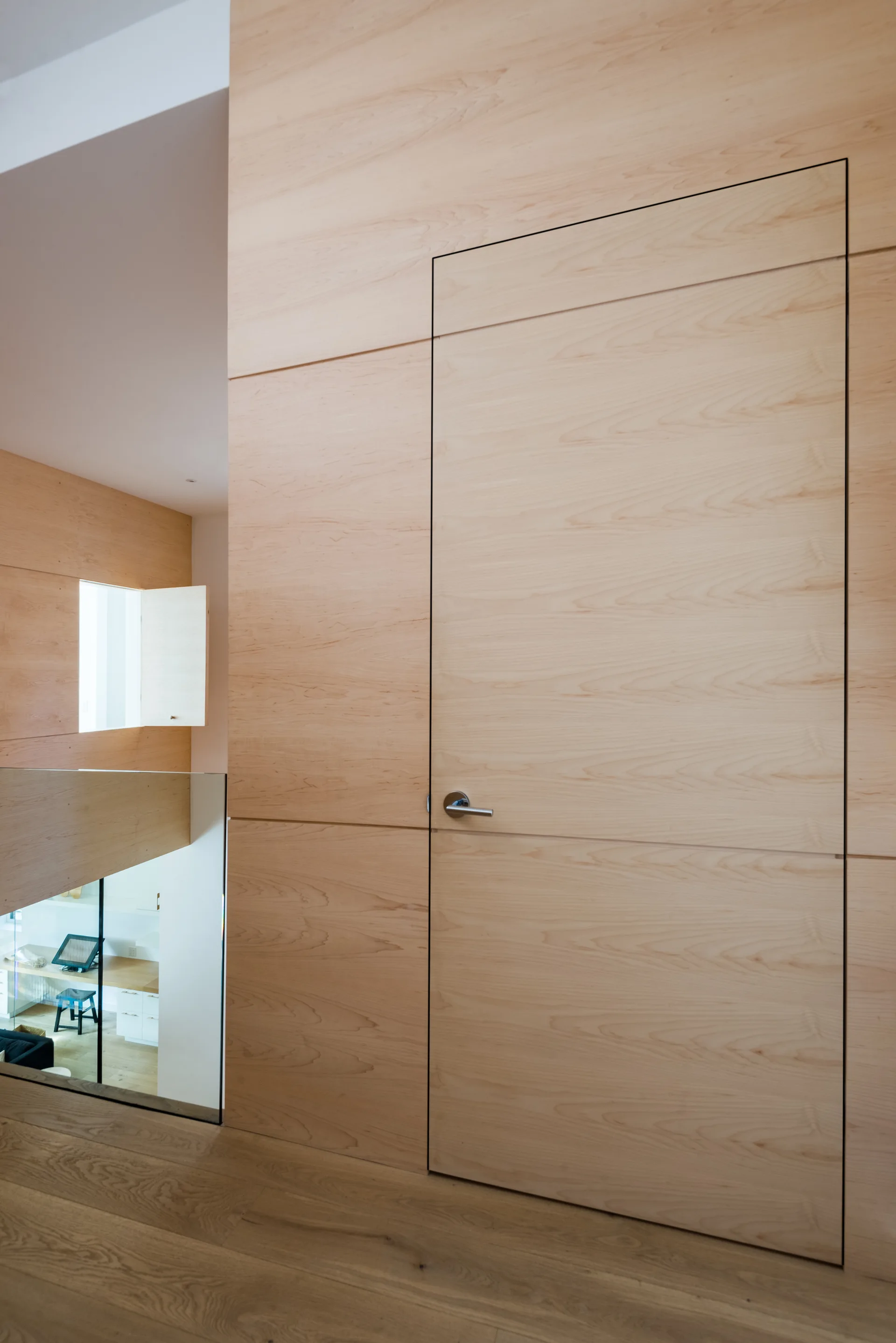 A modern hallway landing features light wood wall paneling with a seamless flush door, a silver handle, and a clear glass railing overlooking the lower level.