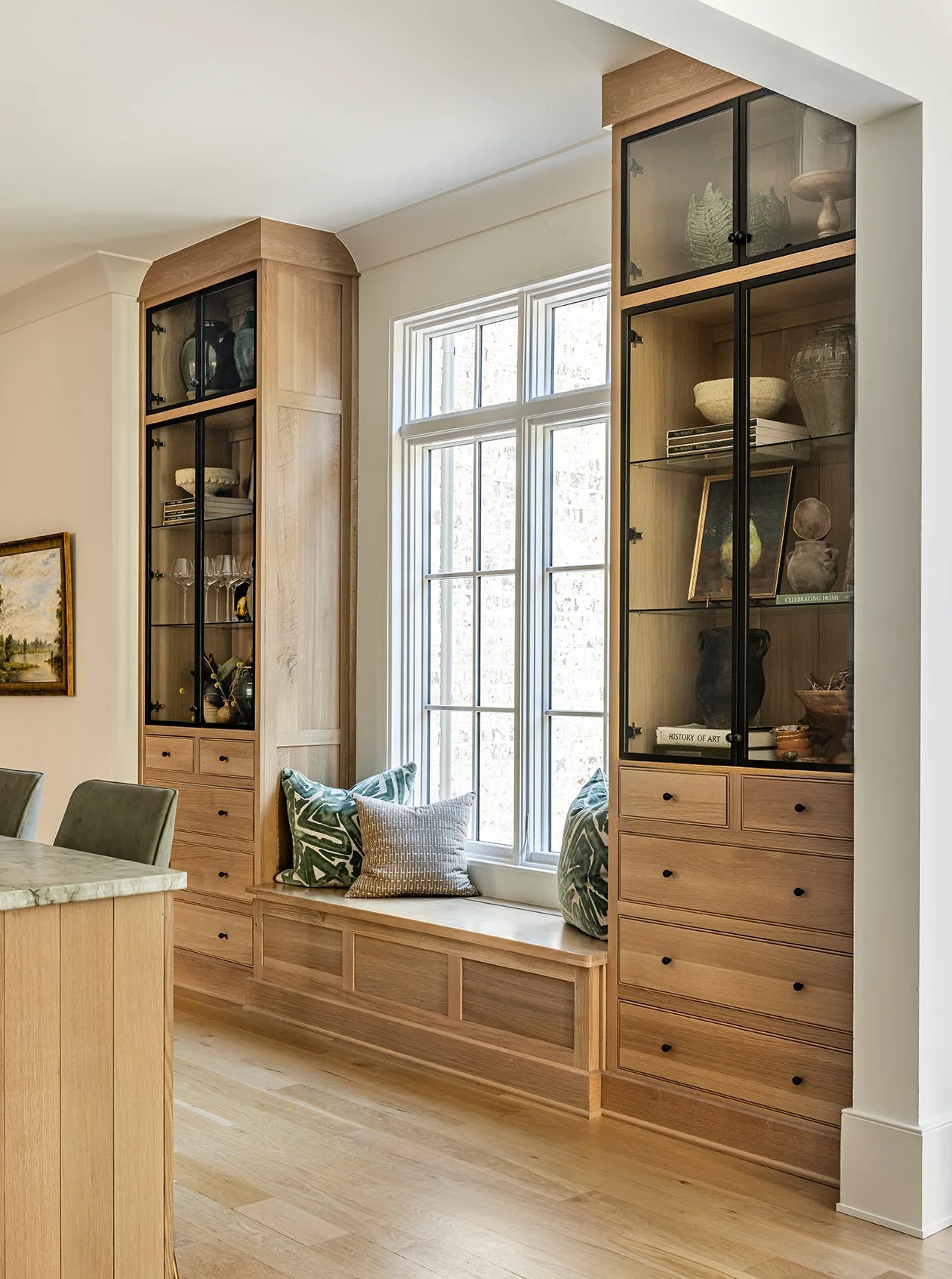 Symmetrical light oak built-in cabinets with glass upper doors and lower drawers flank a window seat with patterned green pillows.