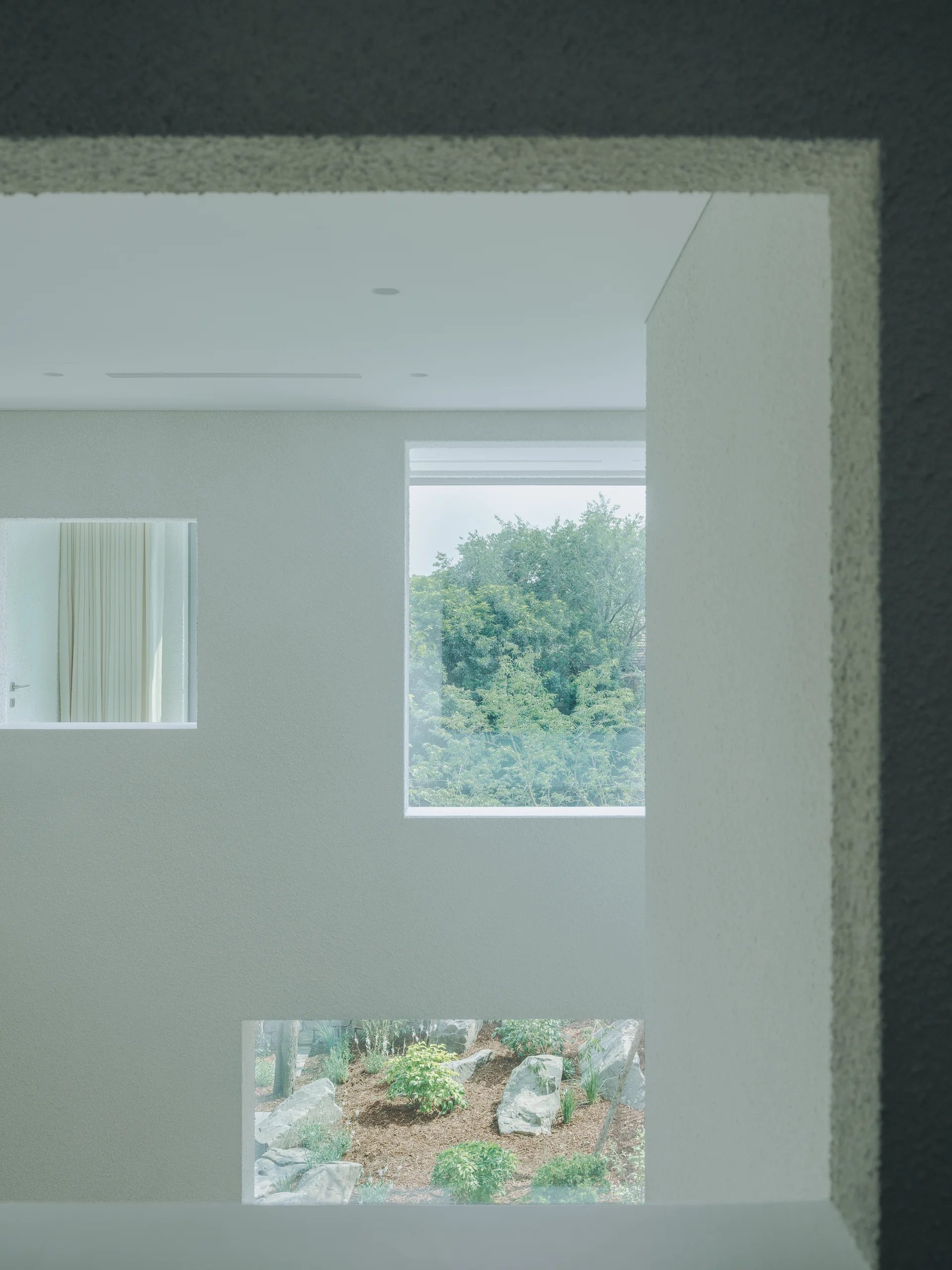 A textured off-white plaster wall features three geometric window openings that frame views of outdoor trees, garden landscaping, and an indoor curtain.