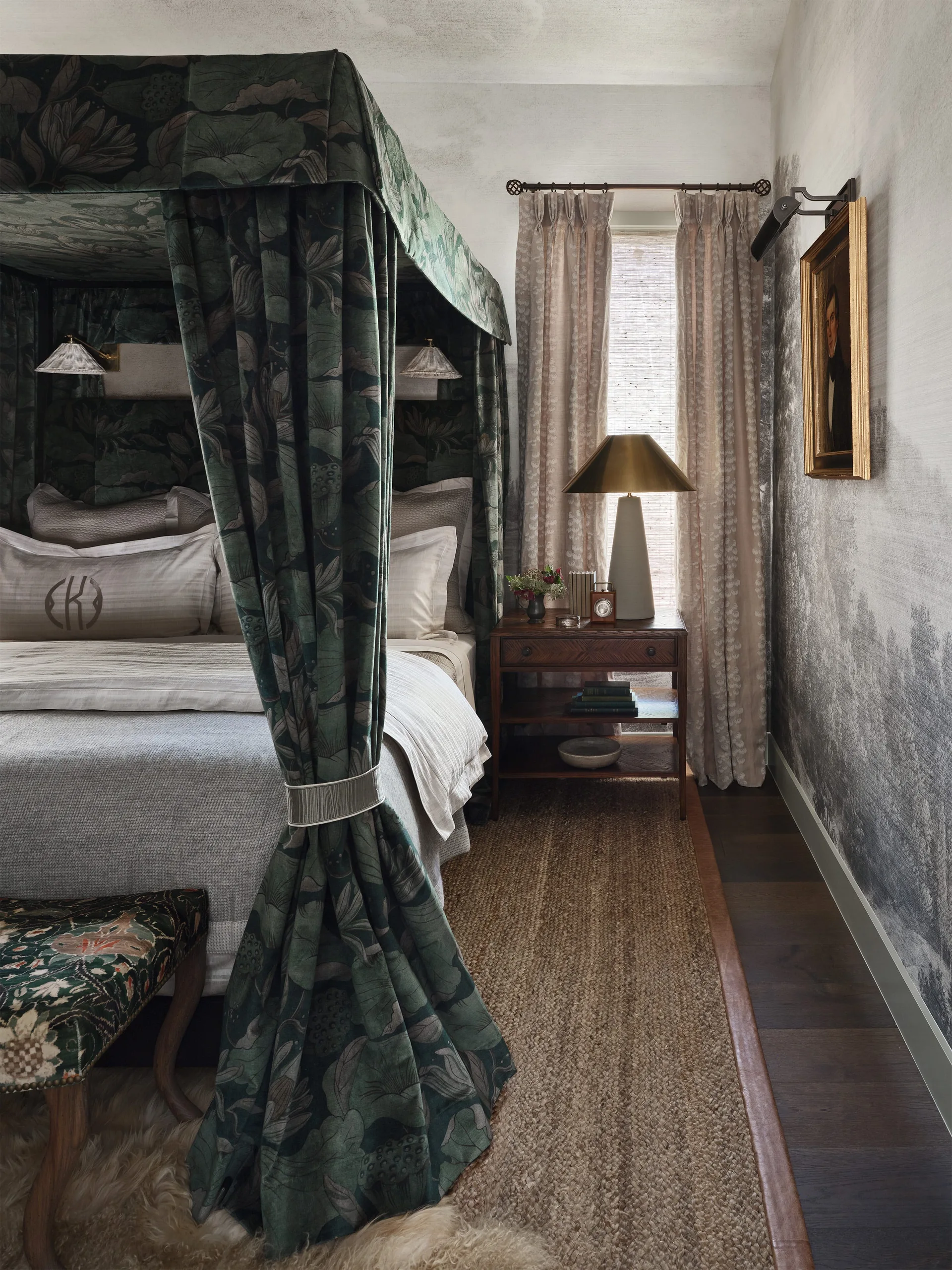 A bedroom features a canopy bed enclosed in green floral fabric, a wooden nightstand with a brass lamp, and grey watercolor-patterned wallpaper.