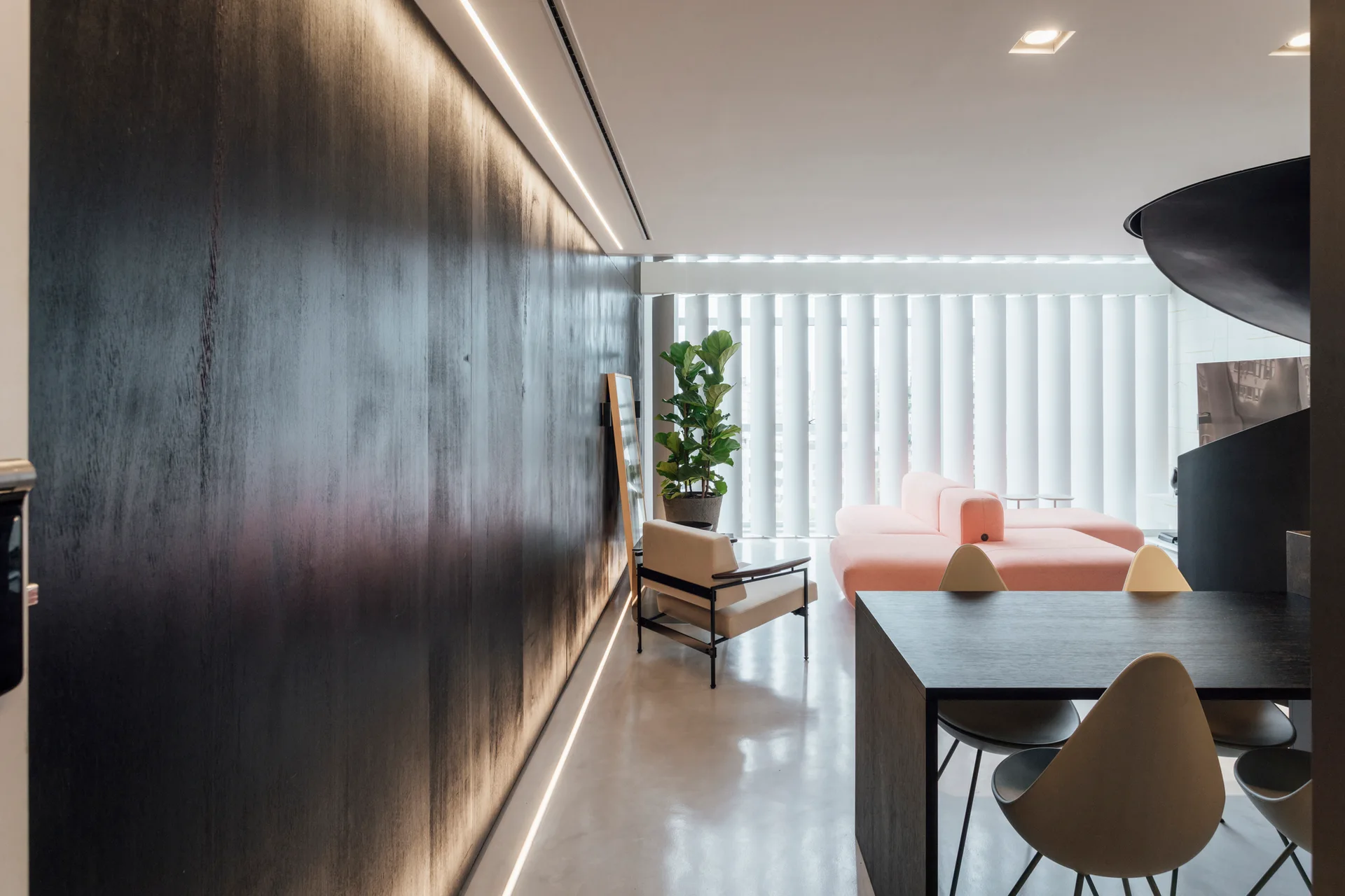 A modern open-concept living area featuring a large illuminated black wall, a low-profile salmon sofa, and a dark wood dining table.
