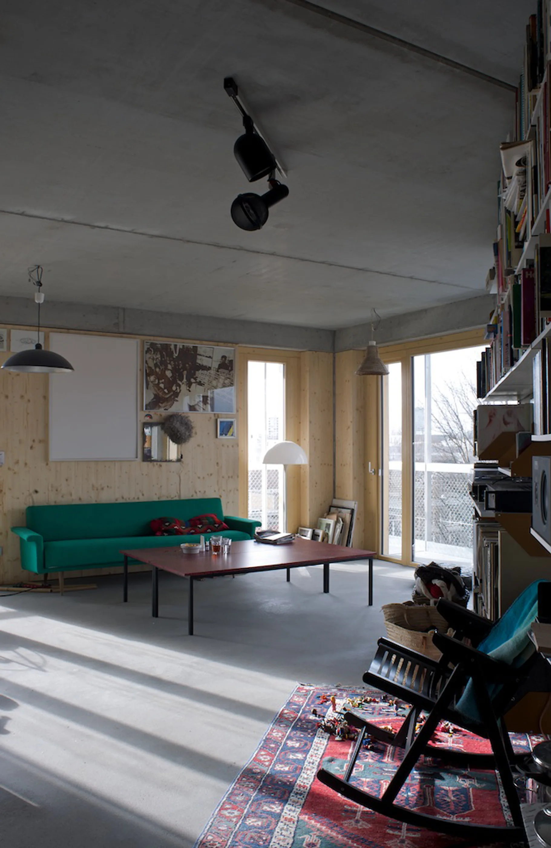 A living room features a bright green sofa against light wood paneled walls, a low red coffee table, and an exposed concrete ceiling.