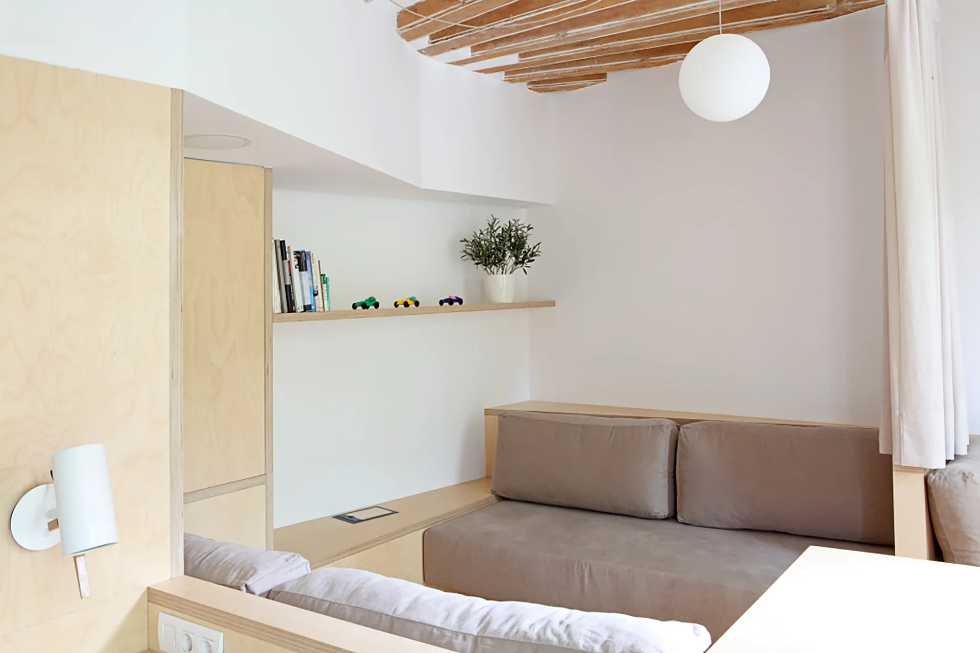 A minimalist living room area featuring a built-in plywood sofa with beige cushions, exposed wooden ceiling rafters, and a white globe pendant light.