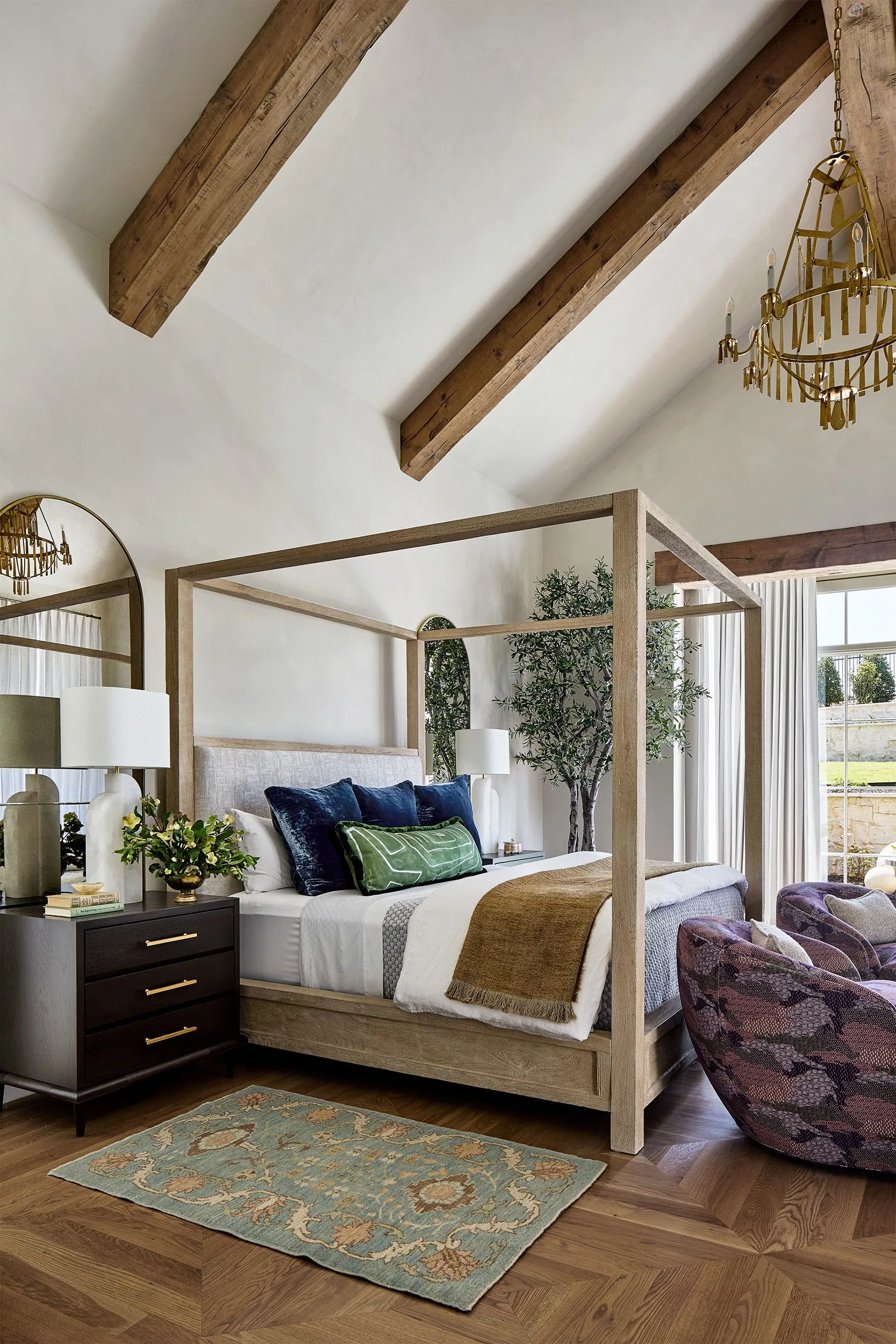 A bedroom features a wood canopy bed, exposed ceiling beams, a dark wood nightstand, and an arched wall mirror.
