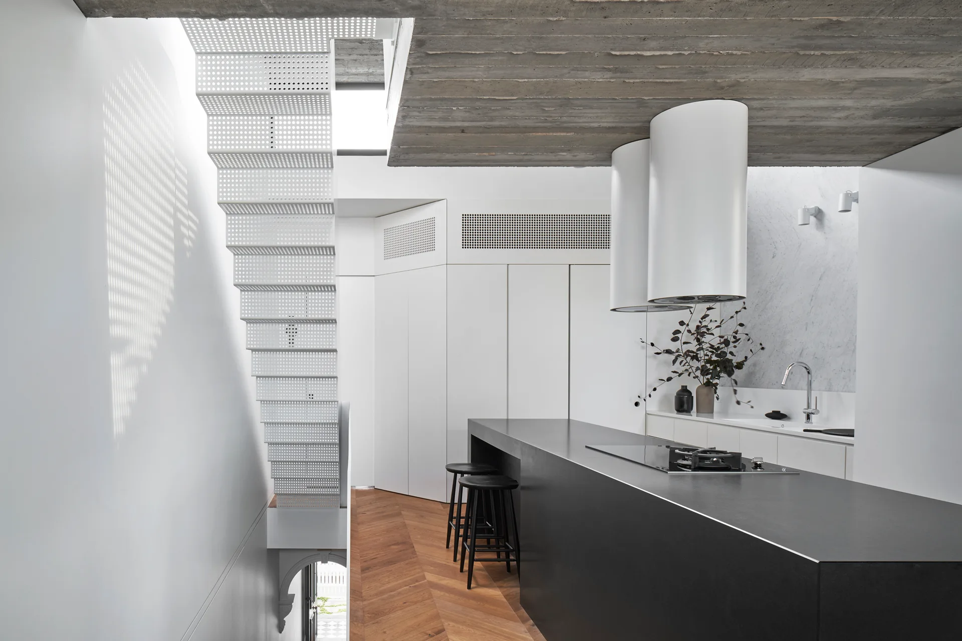 A modern minimalist kitchen features a large black island, two black bar stools, a white perforated metal staircase, and an exposed raw concrete ceiling.