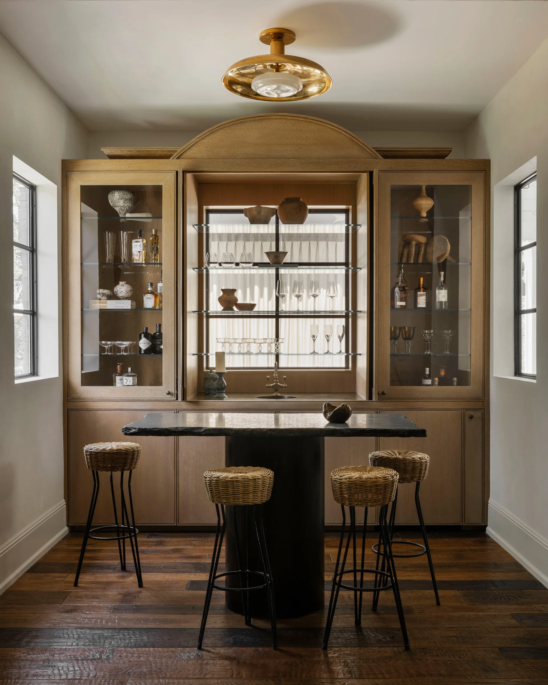 A home bar features a large arched wood cabinet with glass shelves, a black stone island, and four woven rattan stools on dark wood floors.