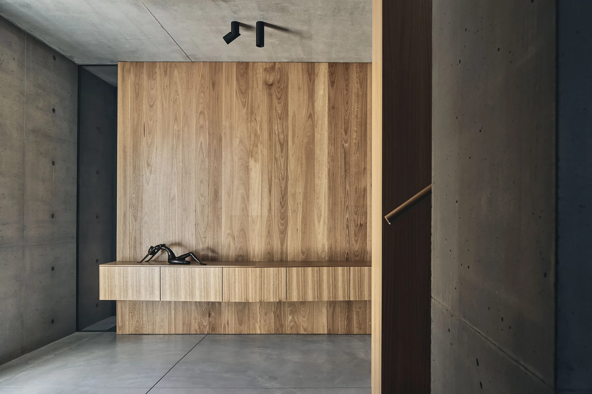 A minimalist entryway features a floating wooden console against a matching wood-paneled wall, set between exposed concrete walls and floor.