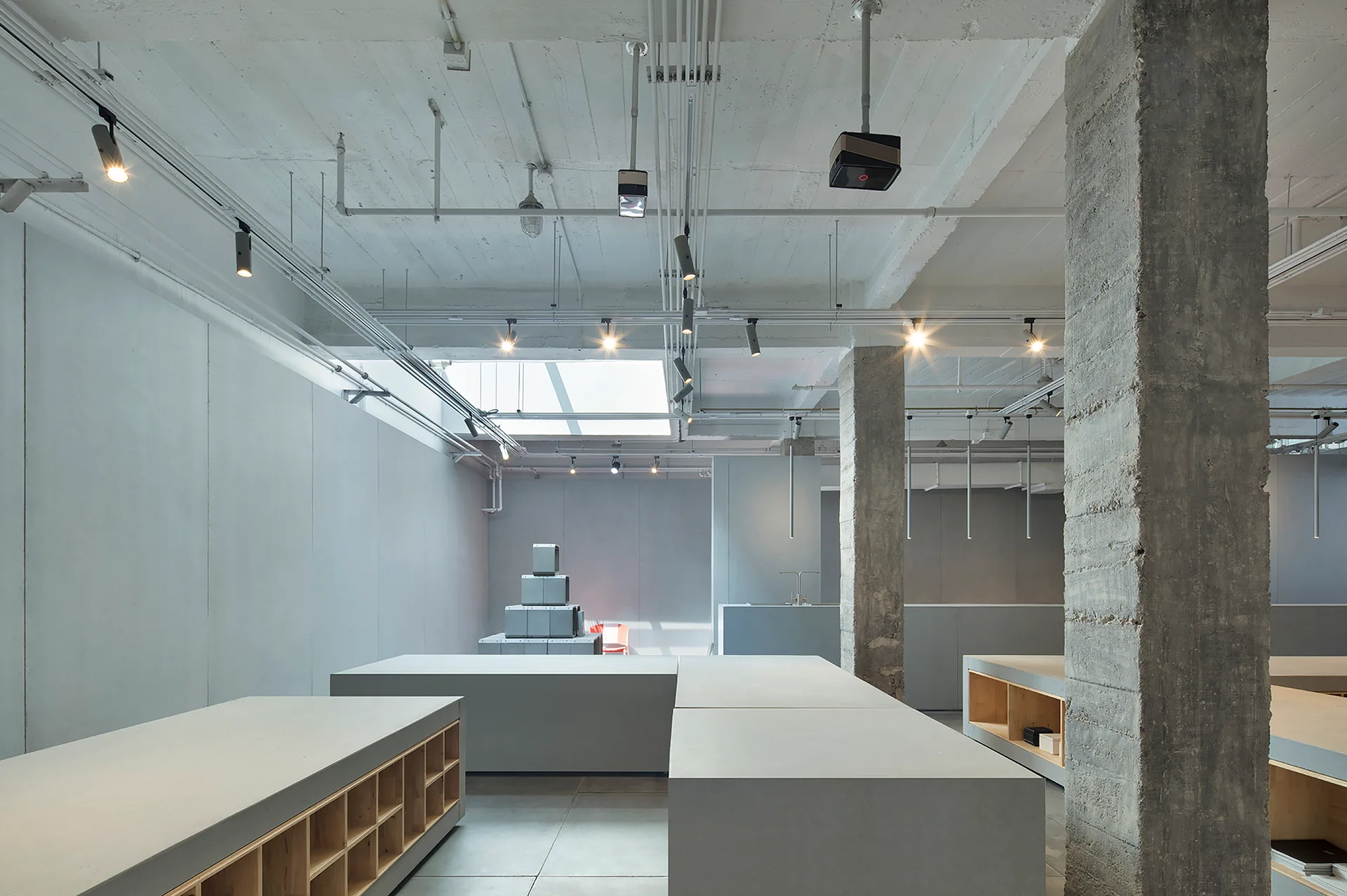 Minimalist grey interior featuring rough concrete pillars, flat display counters with wooden shelving inserts, and exposed track lighting on the ceiling.