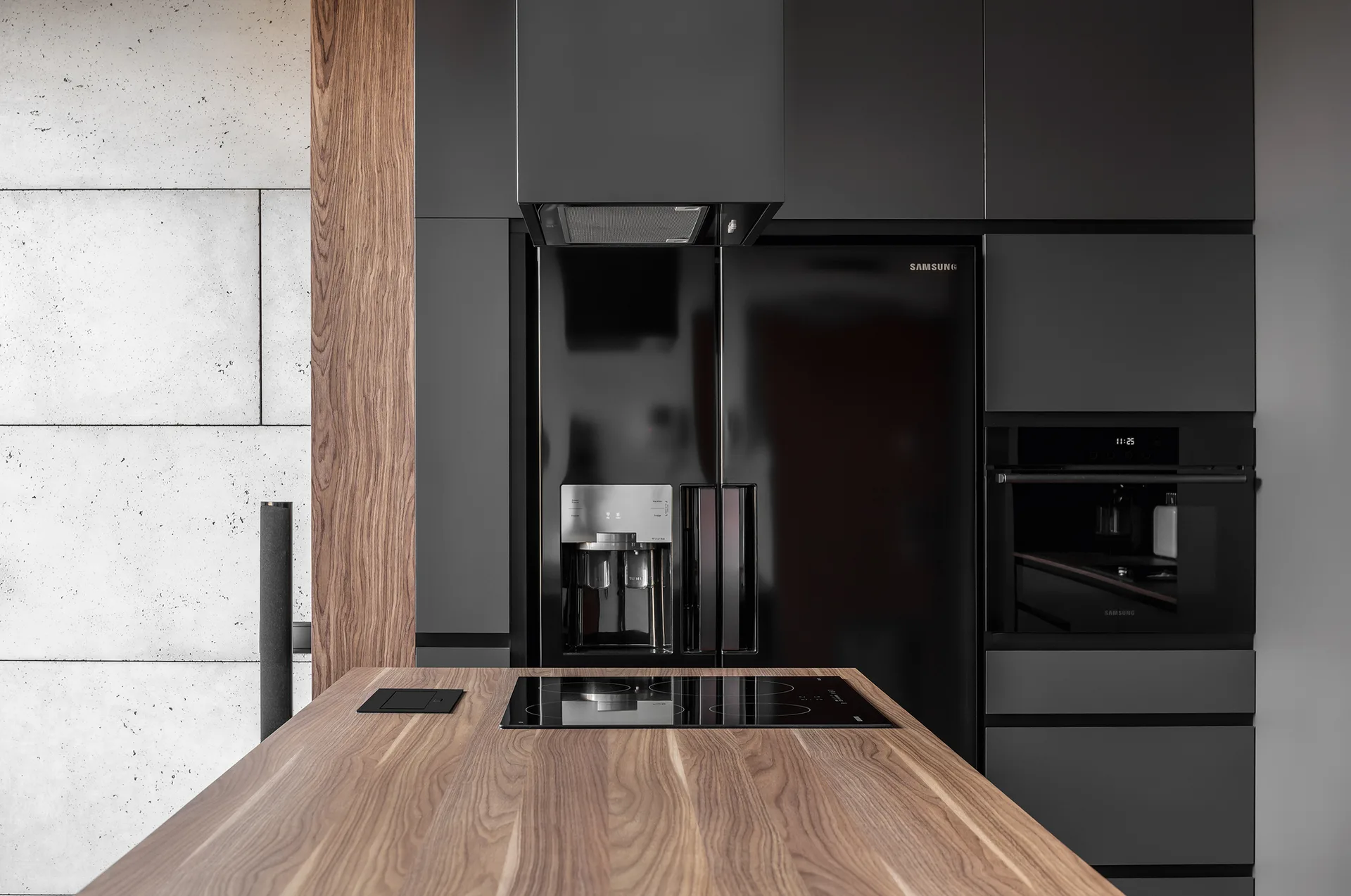 A modern kitchen featuring matte black cabinetry, a wood grain island with an induction cooktop, and concrete-look wall panels.