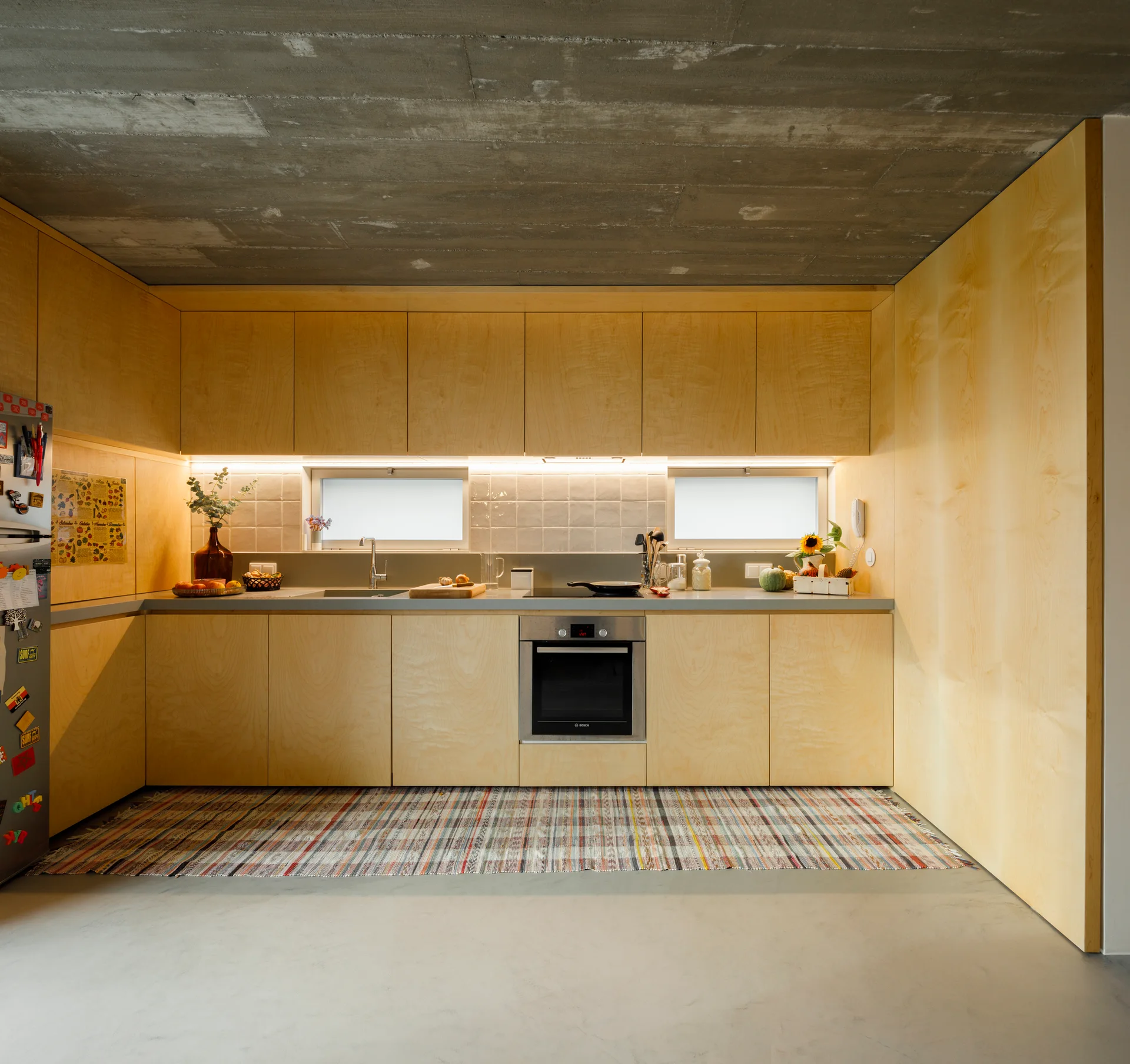 A minimalist kitchen features light wood flat-panel cabinets, an exposed concrete ceiling, a grey countertop, and a striped woven floor rug.