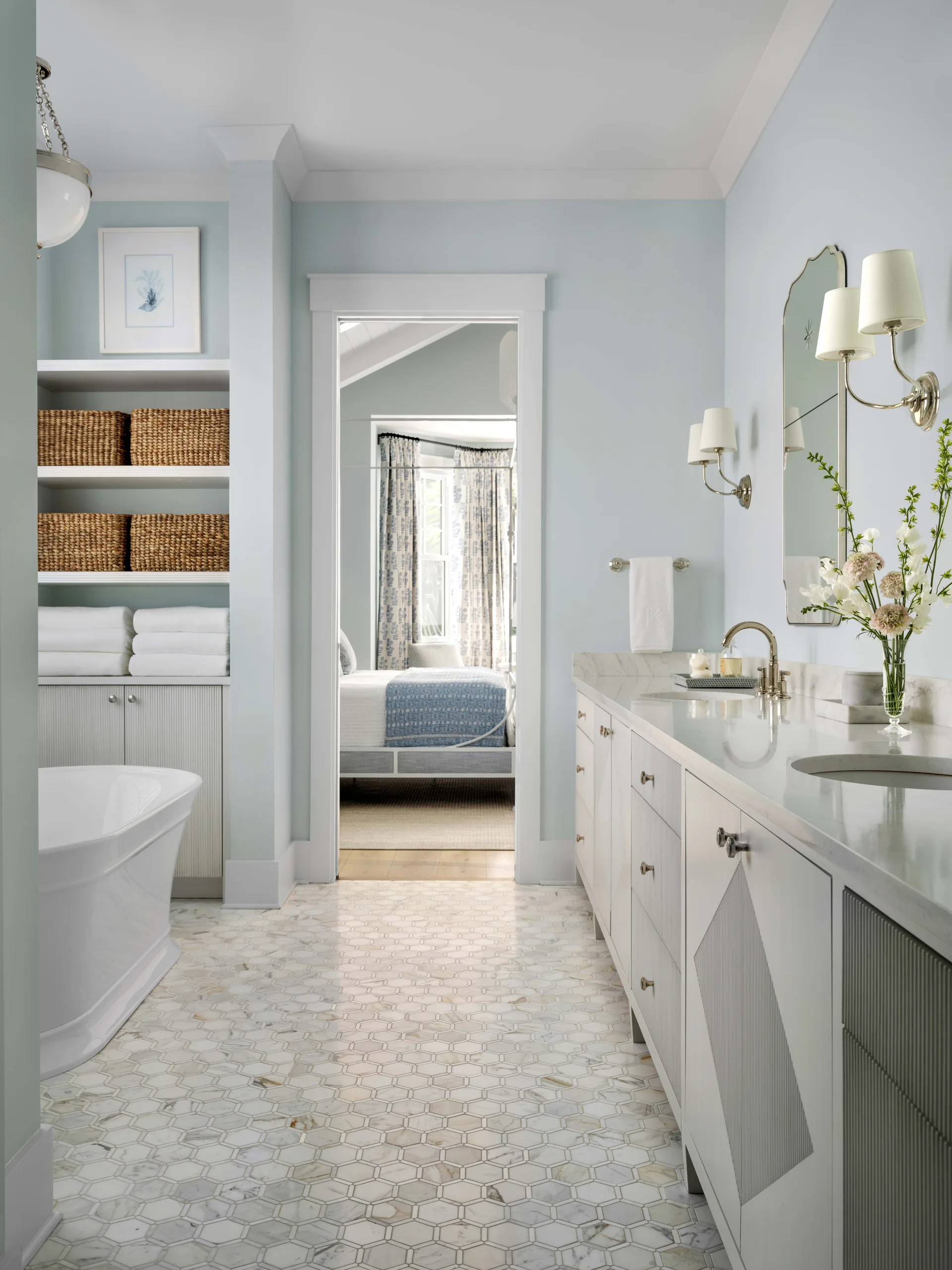 A light blue bathroom features a long white double vanity with marble counters, a freestanding bathtub, and hexagonal marble floor tiles.